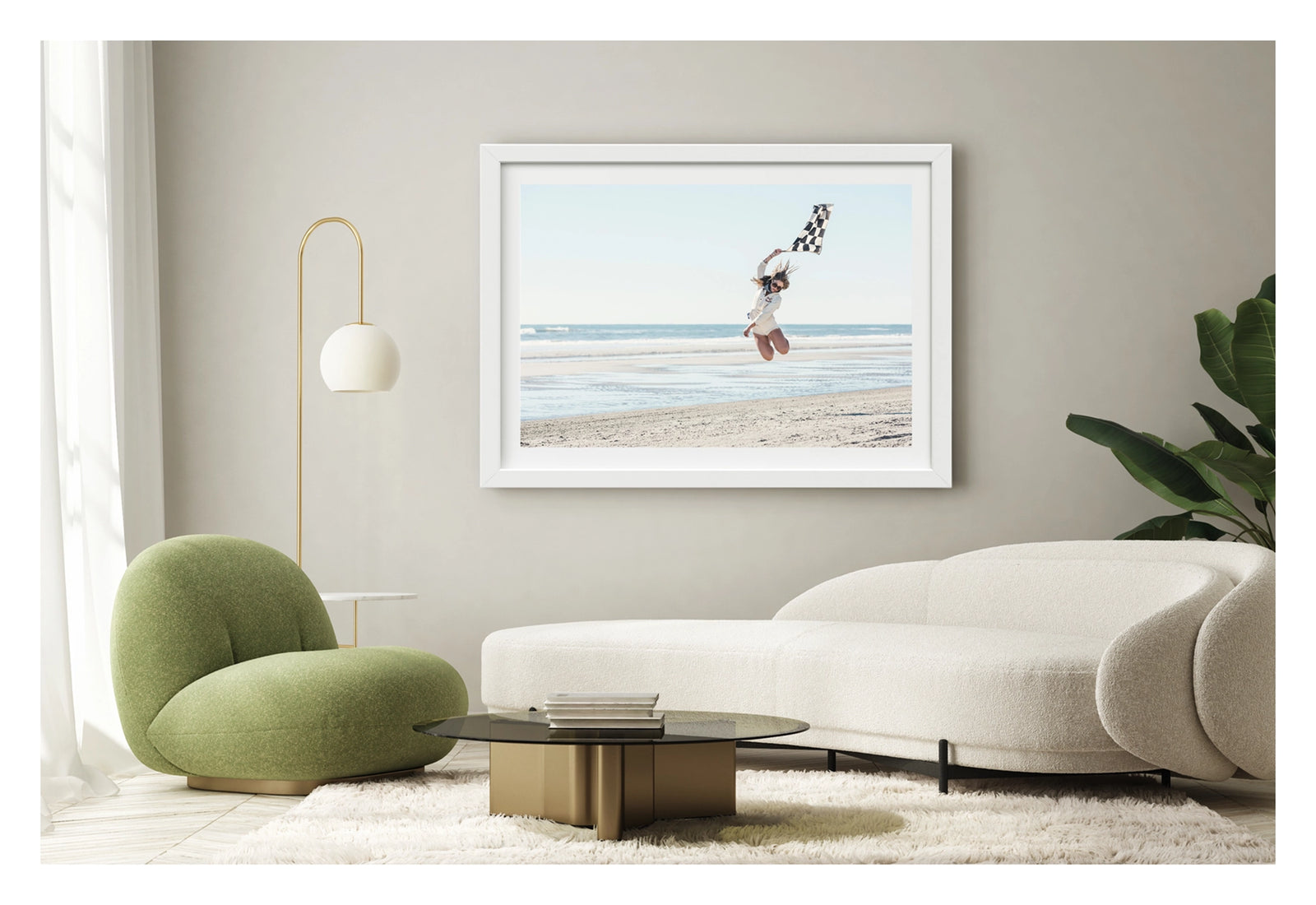 Framed in white, shown in a contemporary living space. A young woman leaps high into the air on a wide empty beach, waving a black and white chequered racing flag above her head as her hair flies in the wind. Wearing a white shirt and shorts, sunglasses and a neck scarf, she is captured mid jump with knees bent against a pale blue sky and gentle surf rolling in behind her. Fine art colour photographic print by Simon Davidson.