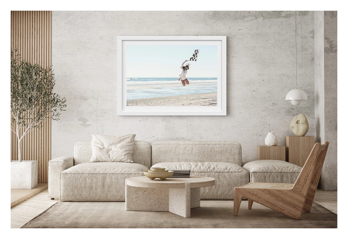 Framed in white, shown in a contemporary living space. A young woman leaps high into the air on a wide empty beach, waving a black and white chequered racing flag above her head as her hair flies in the wind. Wearing a white shirt and shorts, sunglasses and a neck scarf, she is captured mid jump with knees bent against a pale blue sky and gentle surf rolling in behind her. Fine art colour photographic print by Simon Davidson.