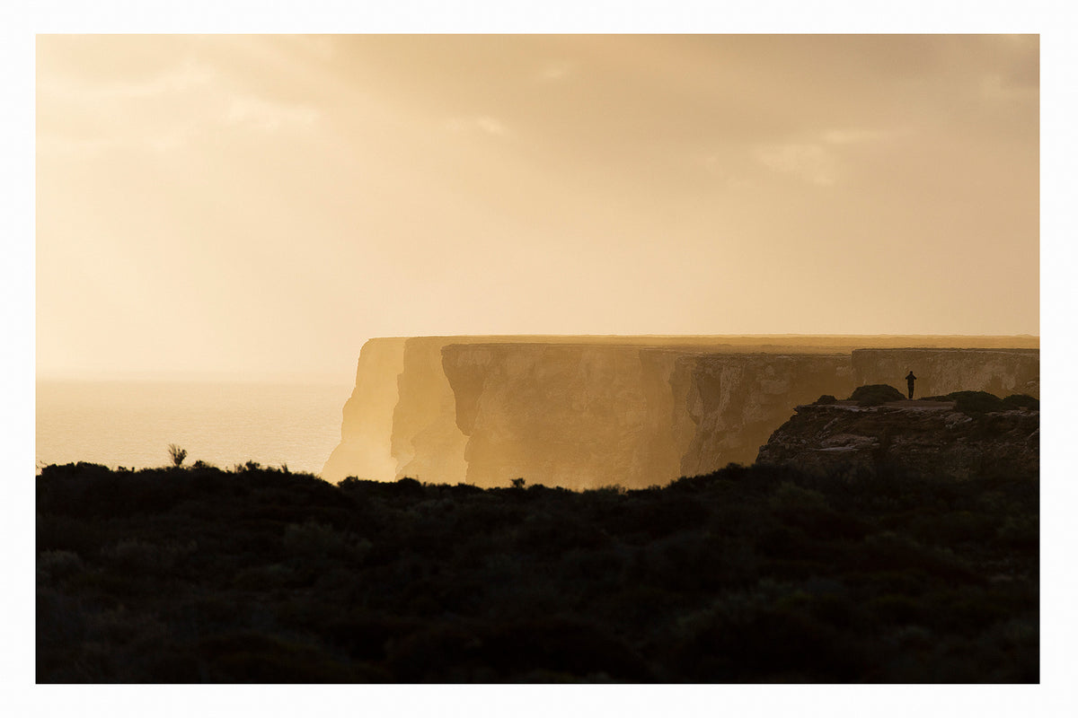In the distance a lone figure stands at the edge of the towering limestone cliffs of the Great Australian Bight, rendered as a tiny silhouette against the warm golden light of dusk. The sheer cliff face stretches across the frame above a hazy Southern Ocean, the dark scrubby foreground in deep shadow while the sky and sea glow in rich amber and gold. Fine art colour photographic print by Simon Davidson.
