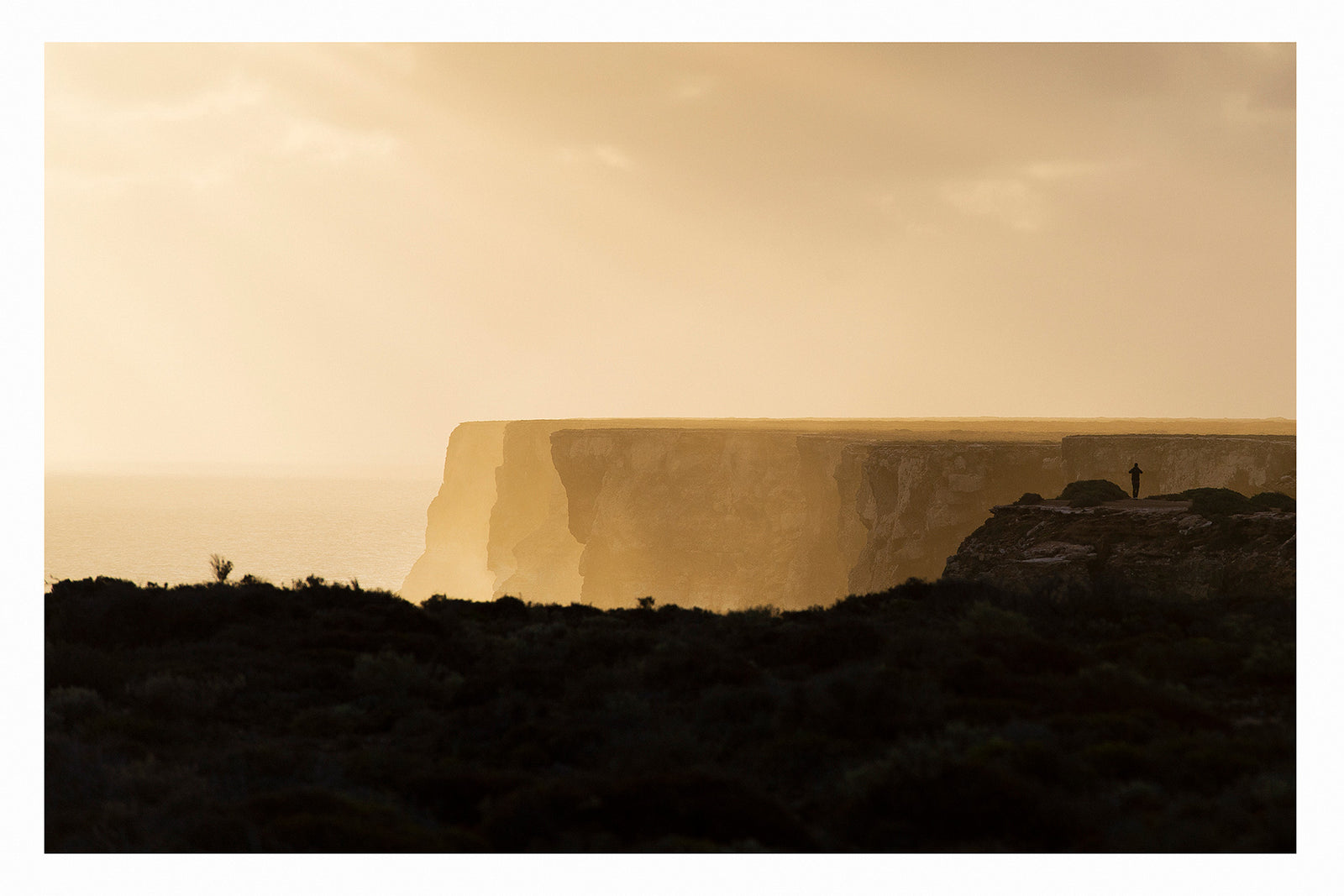In the distance a lone figure stands at the edge of the towering limestone cliffs of the Great Australian Bight, rendered as a tiny silhouette against the warm golden light of dusk. The sheer cliff face stretches across the frame above a hazy Southern Ocean, the dark scrubby foreground in deep shadow while the sky and sea glow in rich amber and gold. Fine art colour photographic print by Simon Davidson.