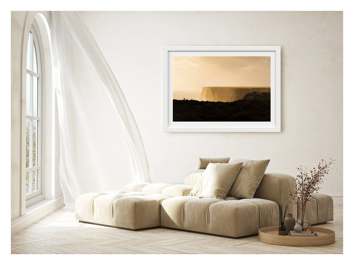 Framed in white, shown in a contemporary living space. We see in the distance a lone figure standing at the edge of the towering limestone cliffs of the Great Australian Bight, rendered as a tiny silhouette against the warm golden light of dusk. The sheer cliff face stretches across the frame above a hazy Southern Ocean, the dark scrubby foreground in deep shadow while the sky and sea glow in rich amber and gold. Fine art colour photographic print by Simon Davidson.
