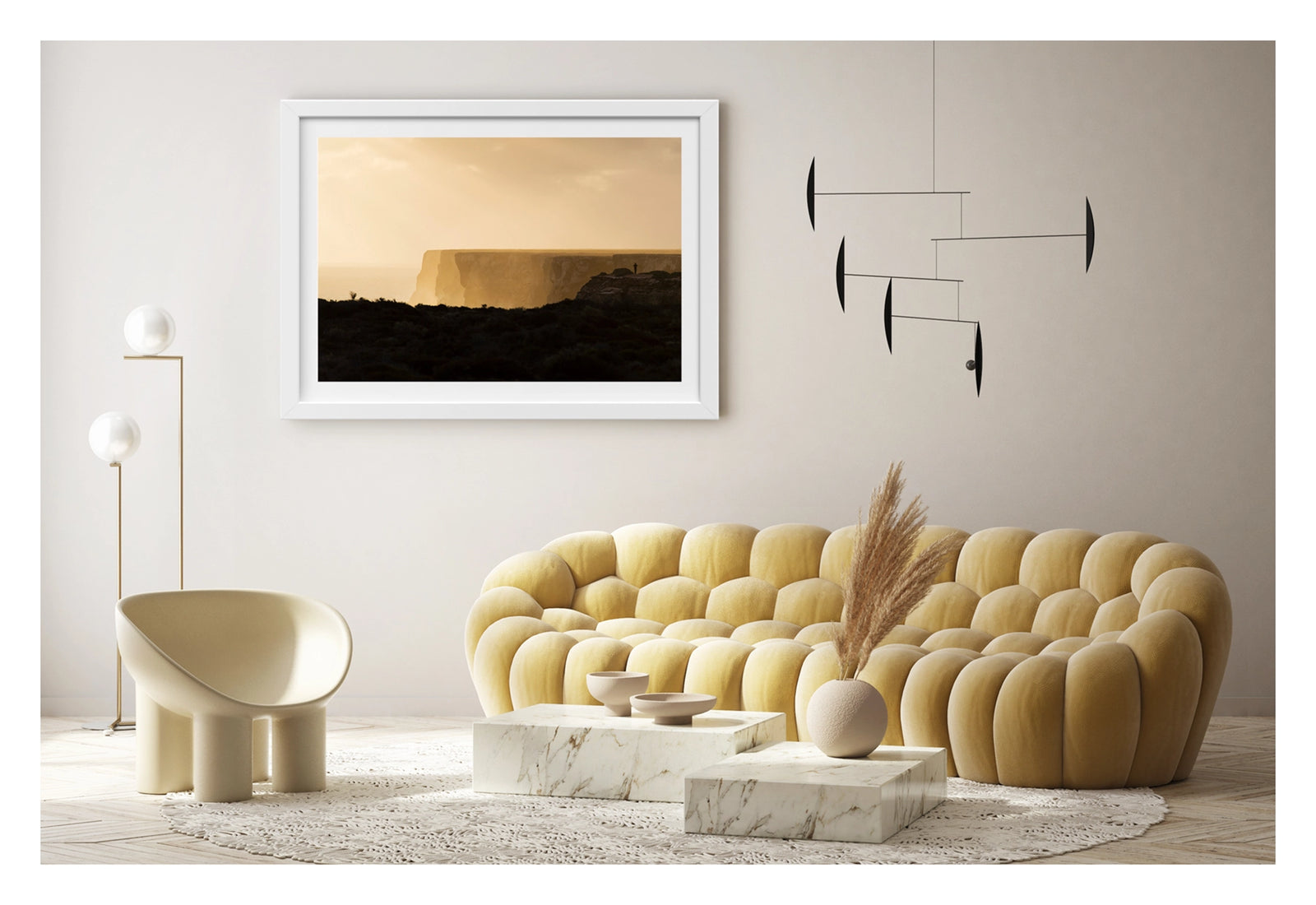 Framed in white, shown in a contemporary living space. We see in the distance a lone figure standing at the edge of the towering limestone cliffs of the Great Australian Bight, rendered as a tiny silhouette against the warm golden light of dusk. The sheer cliff face stretches across the frame above a hazy Southern Ocean, the dark scrubby foreground in deep shadow while the sky and sea glow in rich amber and gold. Fine art colour photographic print by Simon Davidson.