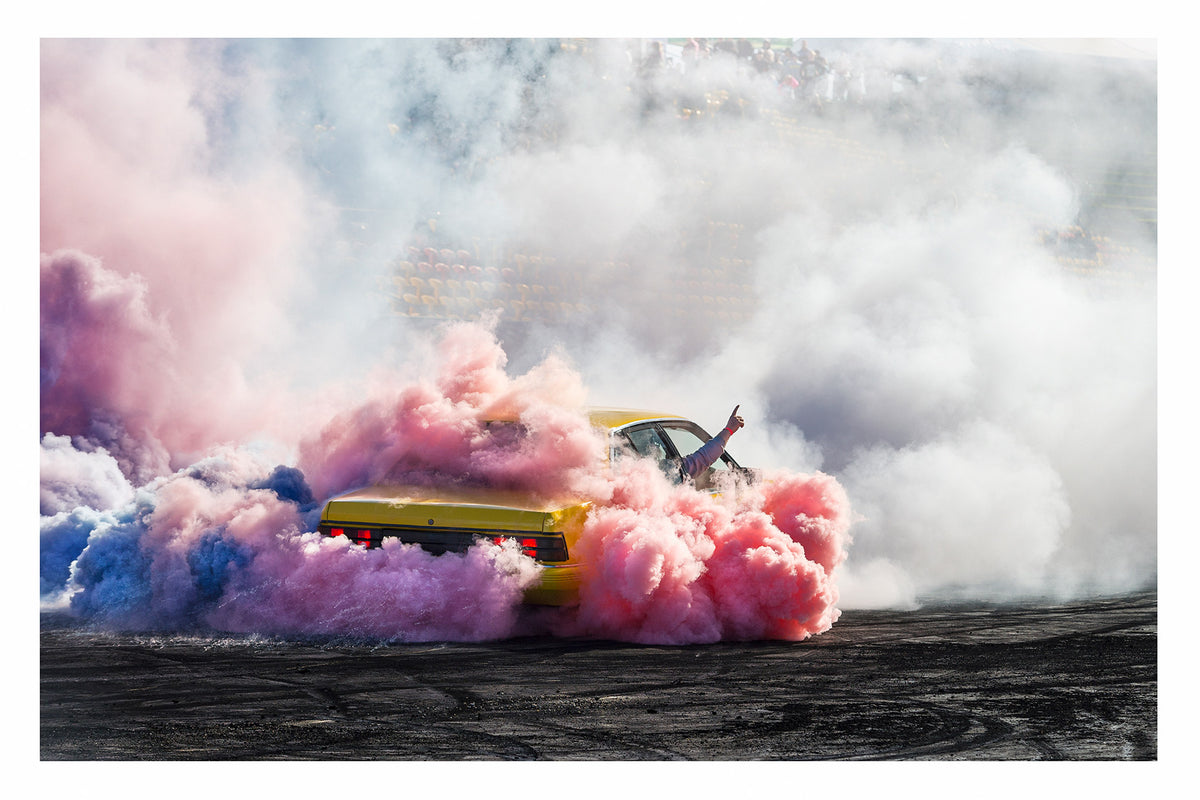 A yellow Holden Commodore performs a burnout before a grandstand crowd, engulfed in a spectacular multicoloured cloud of pink, blue and white tyre smoke that swirls across the tyre marked black bitumen. A driver's arm reaches through the window with a single finger pointed skyward as the vibrant smoke rolls in every direction, the red tail lights glowing through the haze and the blurred faces of the crowd slightly visible through the clearing smoke. Fine art colour photographic print by Simon Davidson.