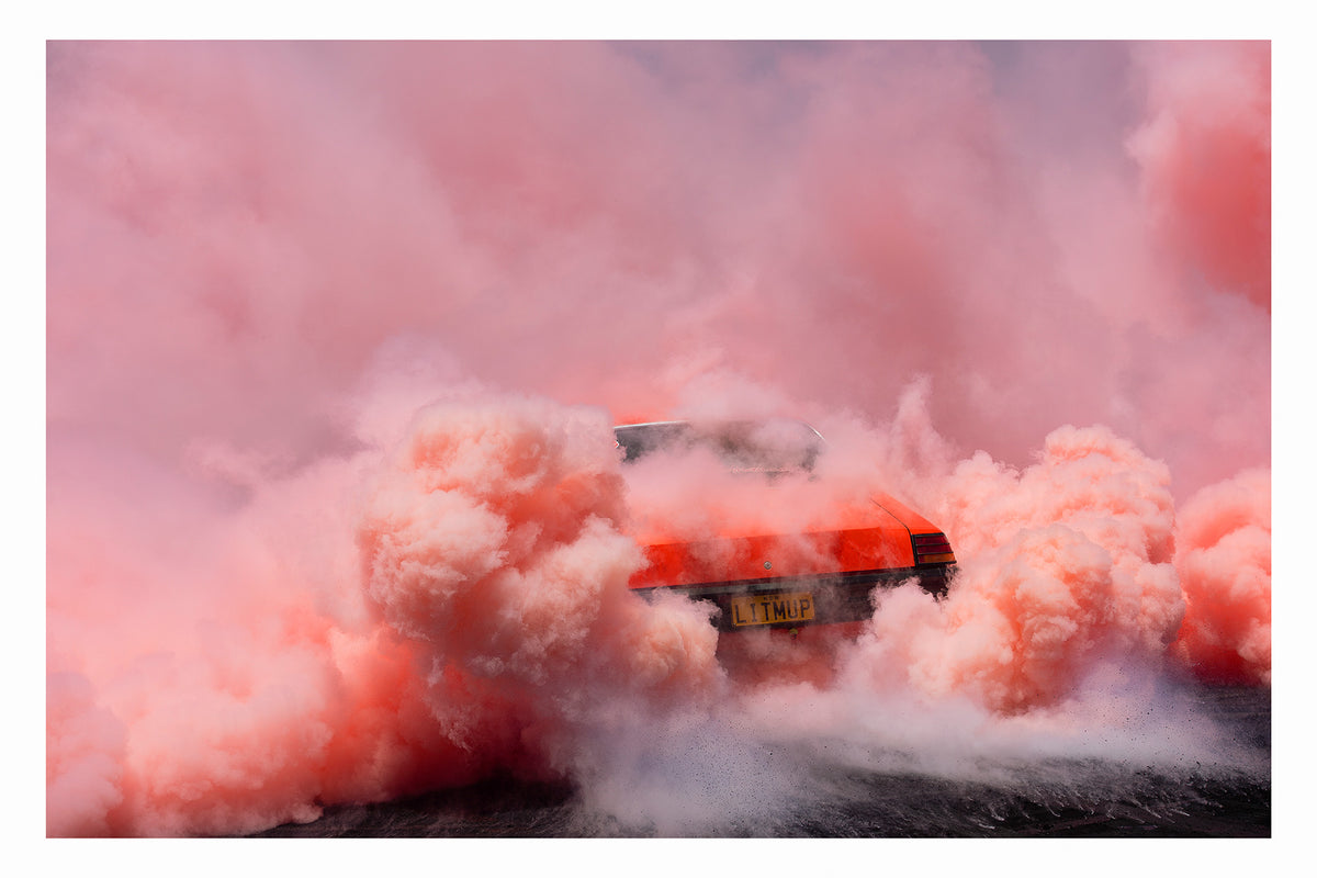 An orange Holden sedan bearing the number plate LITMUP performing a burnout, the car almost entirely consumed by the vast churning cloud of pink/orange tyre smoke that fills the whole frame. The roofline and rear window are just visible through the enveloping haze as pink smoke billows in every direction across the velvety black bitumen below. Fine art colour photographic print by Simon Davidson.