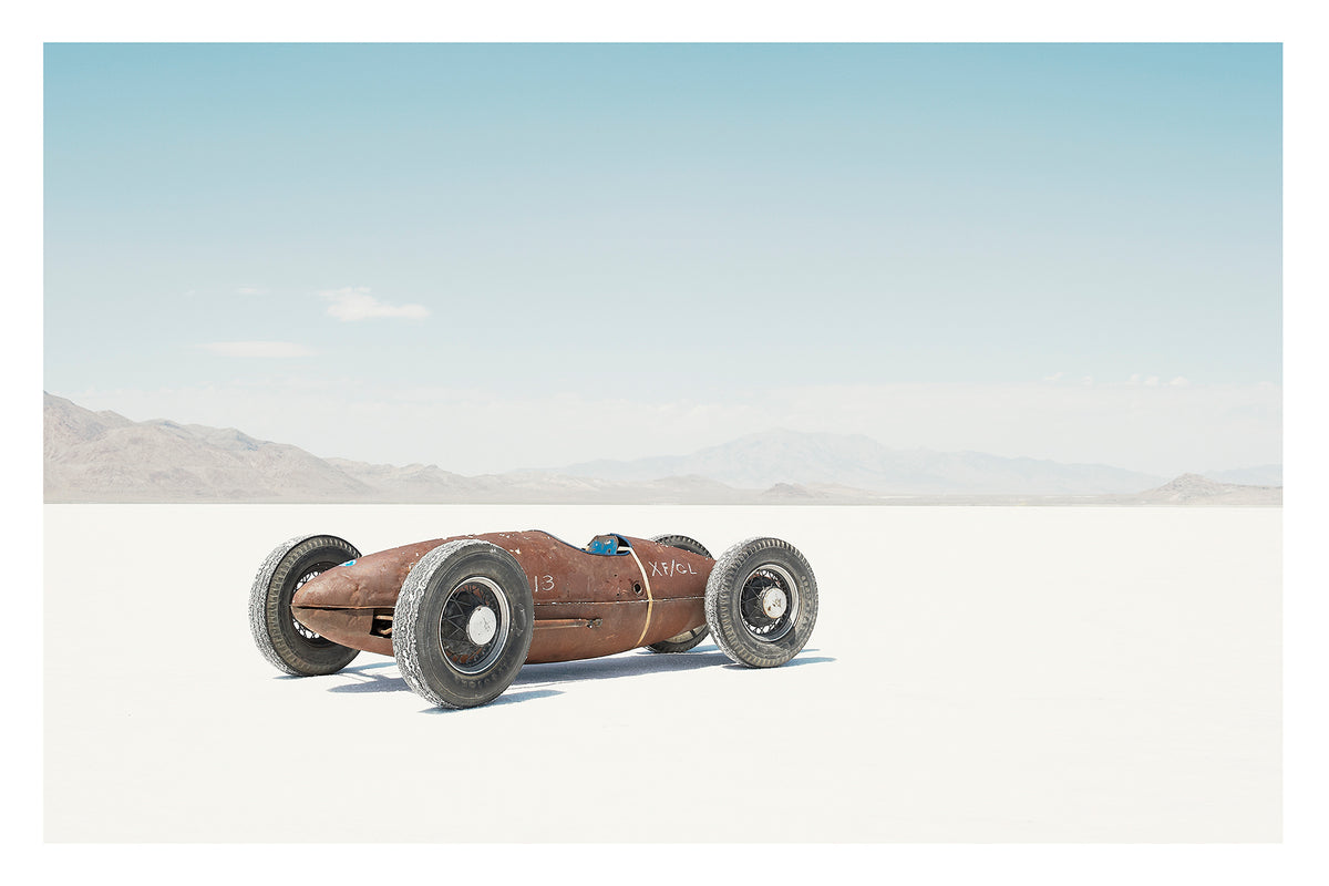 A small rust patinated vintage bellytanker landspeed race car sits alone on the blinding white salt of the Bonneville Salt Flats, its exposed wire wheels and torpedo shaped body giving it the appearance of a miniature prewar racing car. A hazy mountain range dissolves into the pale horizon behind it under a soft blue sky, the tiny machine dwarfed by the vast emptiness surrounding it. Fine art colour photographic print by Simon Davidson.