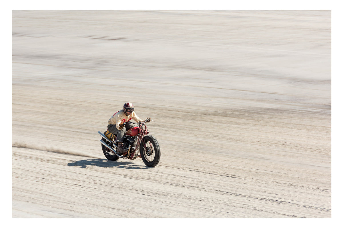 Photographed from above. A motorcycle racer in a cream sweater, goggles and open face helmet leans forward over a red pre-war vintage Harley Davidson as it races down the beach., a small trail of sand kicking up behind as the bike accelerates across the textured surface. Fine art colour photographic print by Simon Davidson.