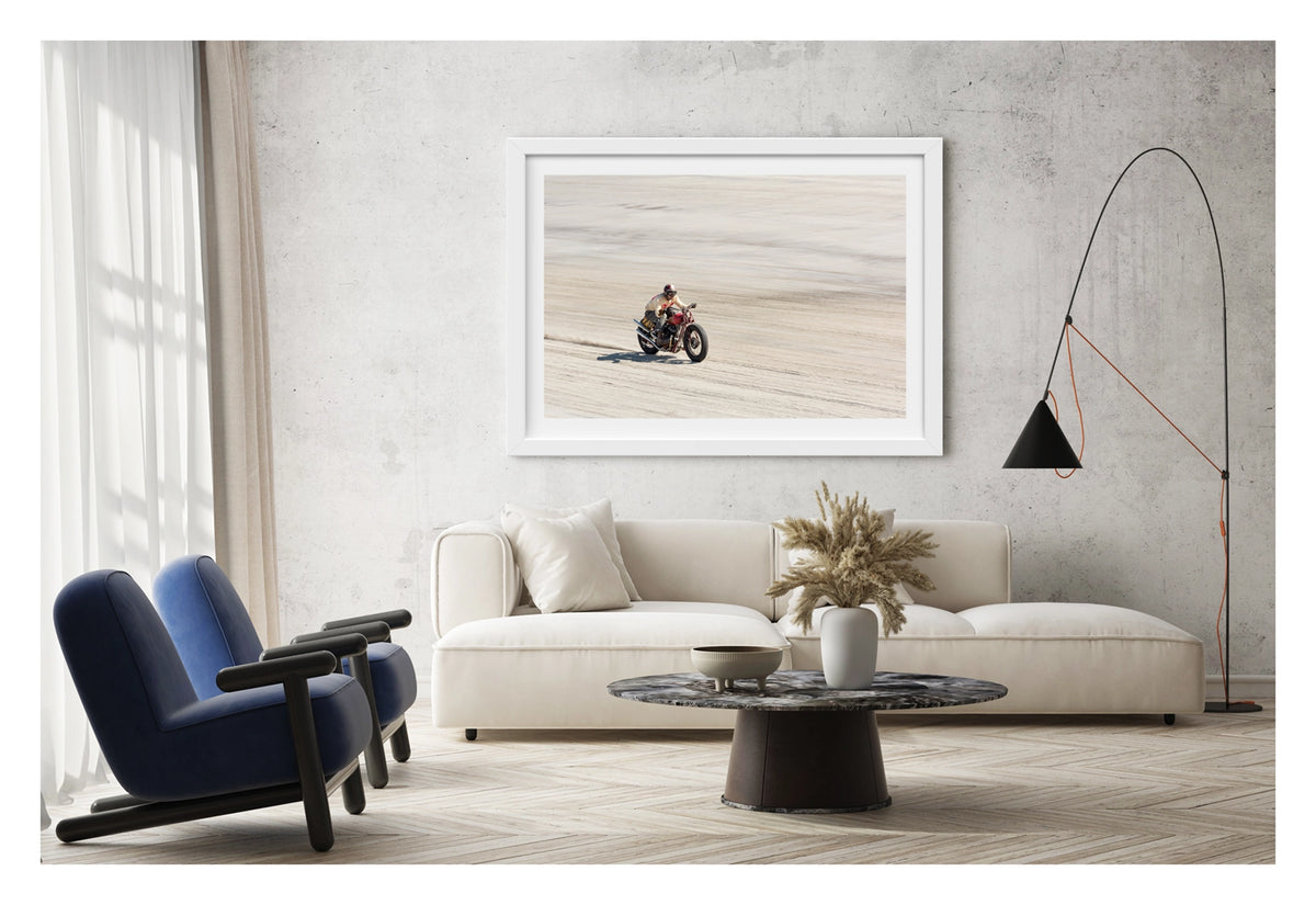 Framed in white, shown in a contemporary space. Photographed from above a motorcycle racer in a cream sweater, goggles and open face helmet leans forward over a red pre-war vintage Harley Davidson as it races down the beach., a small trail of sand kicking up behind as the bike accelerates across the textured surface. Fine art colour photographic print by Simon Davidson.
