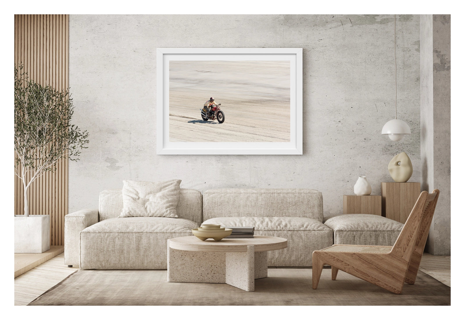 Framed in white, shown in a contemporary space. Photographed from above a motorcycle racer in a cream sweater, goggles and open face helmet leans forward over a red pre-war vintage Harley Davidson as it races down the beach., a small trail of sand kicking up behind as the bike accelerates across the textured surface. Fine art colour photographic print by Simon Davidson.