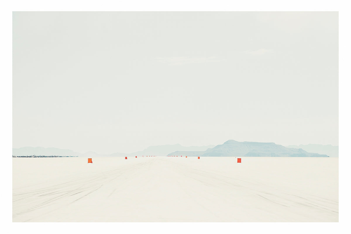 A row of small orange course markers stretches into the vanishing point across the blinding white surface of the Bonneville Salt Flats, tyre tracks scoring the salt in the foreground as distant mountains dissolve into a pale grey horizon. The vast emptiness of the scene and the near seamless merging of salt flat and sky give the image a stark, minimalist beauty. Fine art colour photographic print by Simon Davidson.