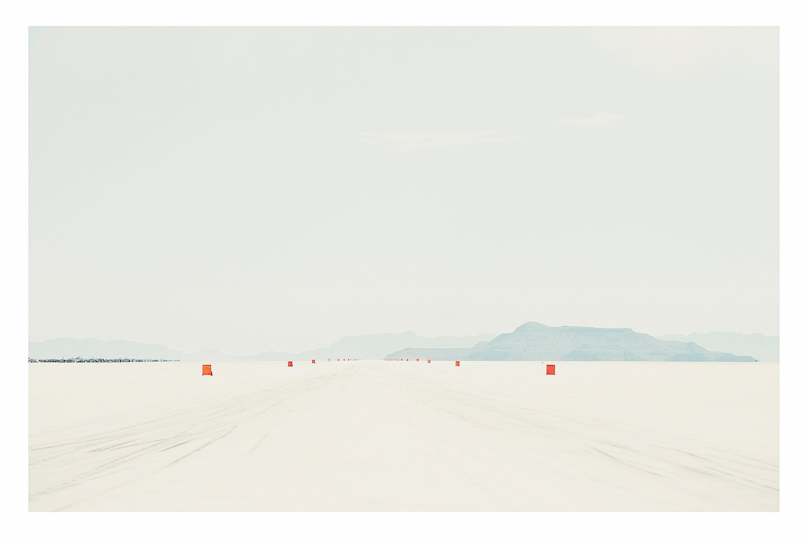 A row of small orange course markers stretches into the vanishing point across the blinding white surface of the Bonneville Salt Flats, tyre tracks scoring the salt in the foreground as distant mountains dissolve into a pale grey horizon. The vast emptiness of the scene and the near seamless merging of salt flat and sky give the image a stark, minimalist beauty. Fine art colour photographic print by Simon Davidson.