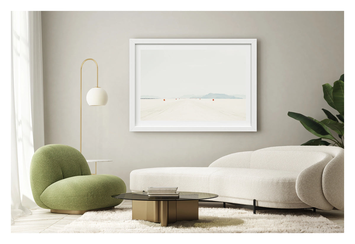 Framed in white, shown in a contemporary living space. We see a row of small orange course markers stretches into the vanishing point across the blinding white surface of the Bonneville Salt Flats, tyre tracks scoring the salt in the foreground as distant mountains dissolve into a pale grey horizon. The vast emptiness of the scene and the near seamless merging of salt flat and sky give the image a stark, minimalist beauty. Fine art colour photographic print by Simon Davidson.