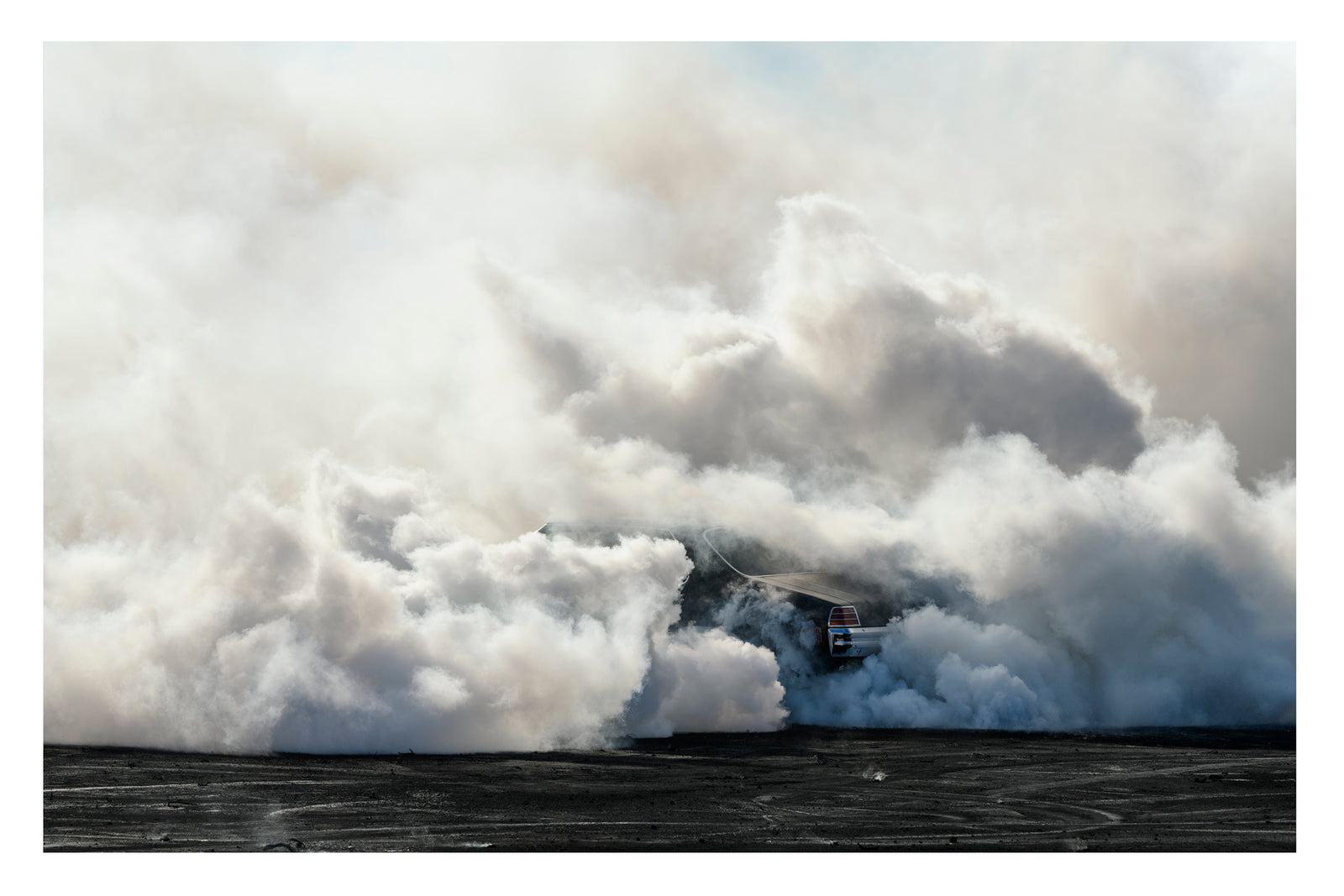 We see the rear of a black Holden GTS sedan is almost entirely swallowed by a towering cloud of white tyre smoke during a burnout, with only the red tail light and rear chrome bumper visible through the billowing haze. The smoke builds into a massive marshmallow like formation that fills the frame from the black bitumen cove red in burnout detritus to the pale sky above, dwarfing the car beneath it. Fine art colour photographic print by Simon Davidson.
