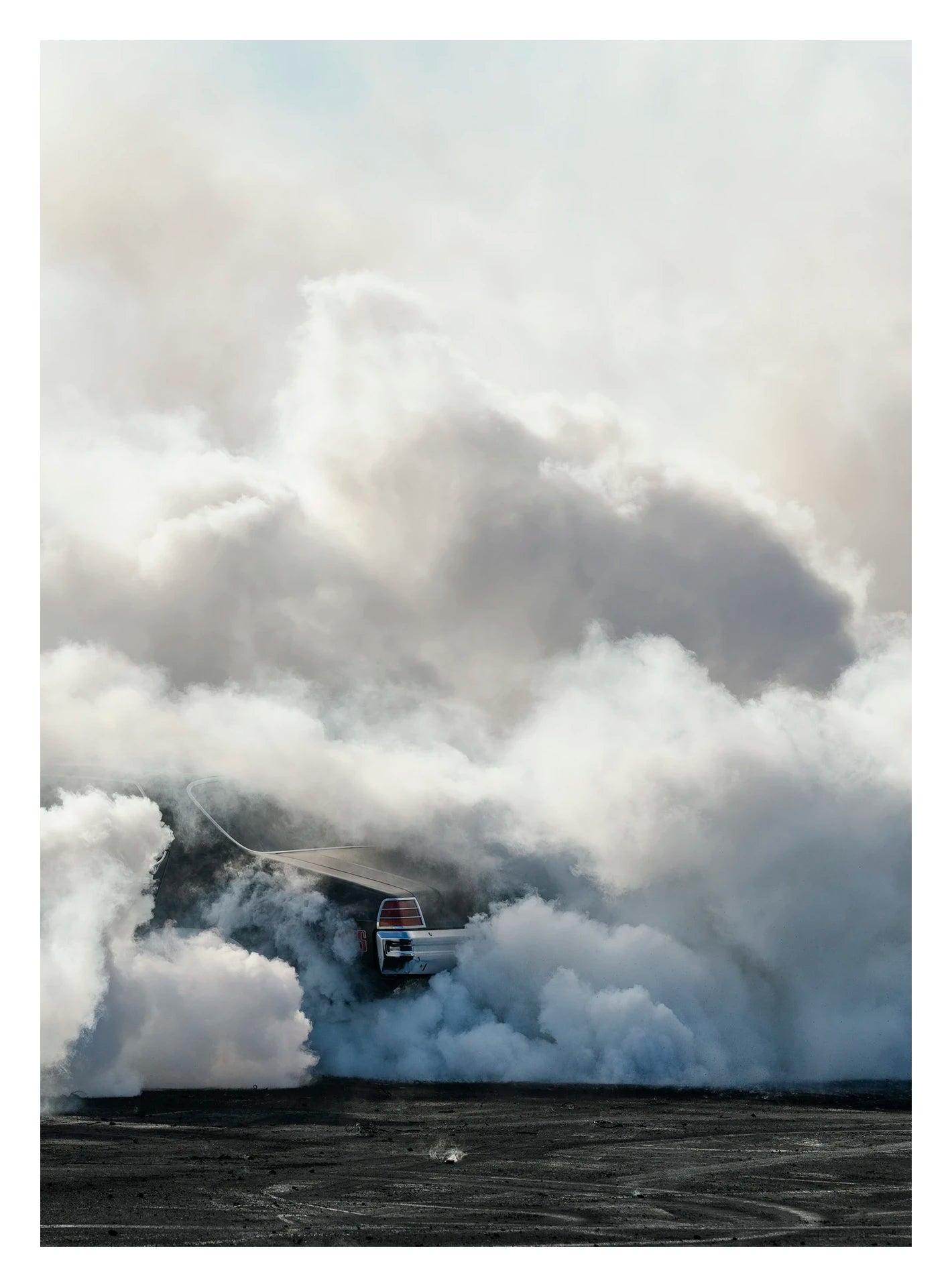 The rear of a black Holden GTS sedan is almost entirely swallowed by a towering cloud of white tyre smoke during a burnout, with only the red tail light and rear chrome bumper visible through the billowing haze. The smoke builds into a massive marshmallow like formation that fills the frame from the black bitumen covered in burnout detritus to the pale sky above, dwarfing the car beneath it. Fine art colour photographic print by Simon Davidson.
