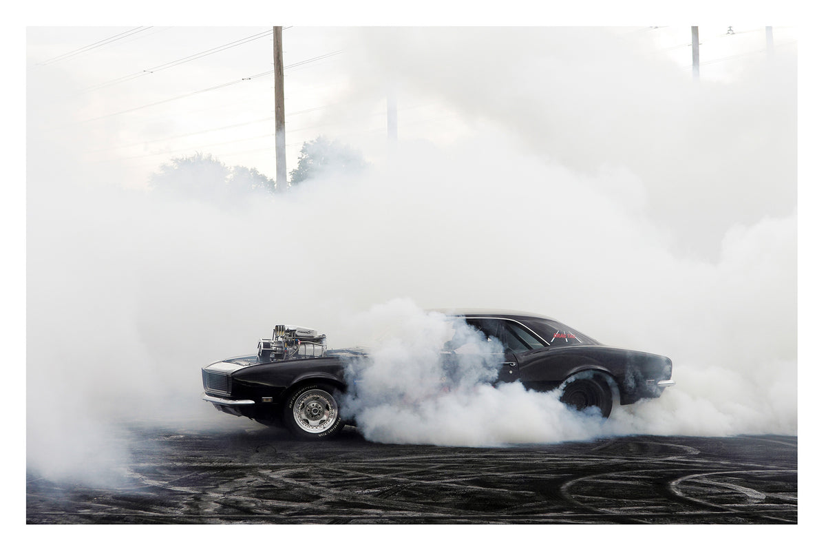 A black Chevrolet Camaro with a polished supercharger protruding through the bonnet performs a burnout on a rubber streaked bitumen surface, engulfed in a thick rolling cloud of white tyre smoke that erases the sky. Power lines and trees ghost through the haze in the background as the car sits at the centre of its own self made storm, wide slick tyres barely visible beneath the billowing smoke. Fine art colour photographic print by Simon Davidson.