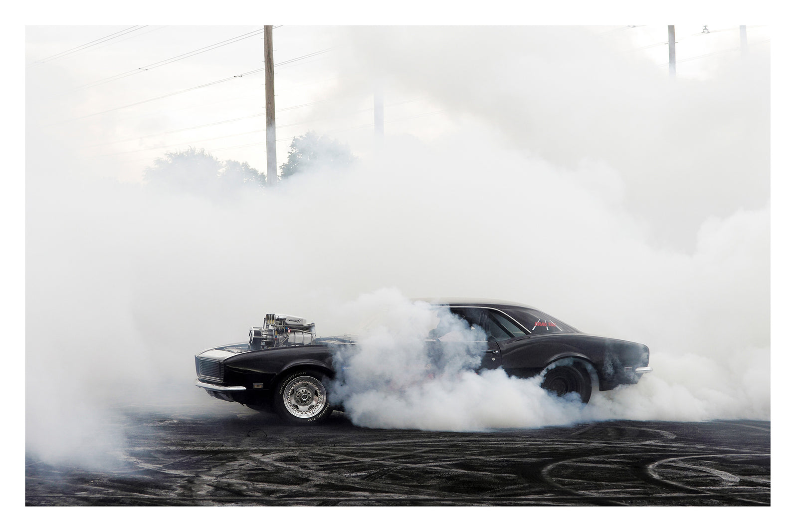 A black Chevrolet Camaro with a polished supercharger protruding through the bonnet performs a burnout on a rubber streaked bitumen surface, engulfed in a thick rolling cloud of white tyre smoke that erases the sky. Power lines and trees ghost through the haze in the background as the car sits at the centre of its own self made storm, wide slick tyres barely visible beneath the billowing smoke. Fine art colour photographic print by Simon Davidson.