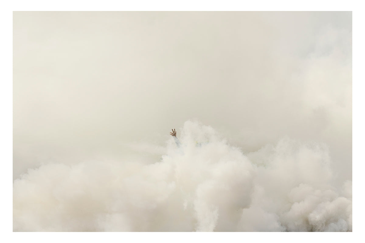 A single human hand reaches up from a vast rolling sea of white tyre smoke, fingers splayed open against a pale hazy sky, the rest of the body completely swallowed by the billowing cloud below. The minimalist composition and vast expanse of white smoke give the image an almost surreal, painterly quality. Fine art colour photographic print by Simon Davidson.