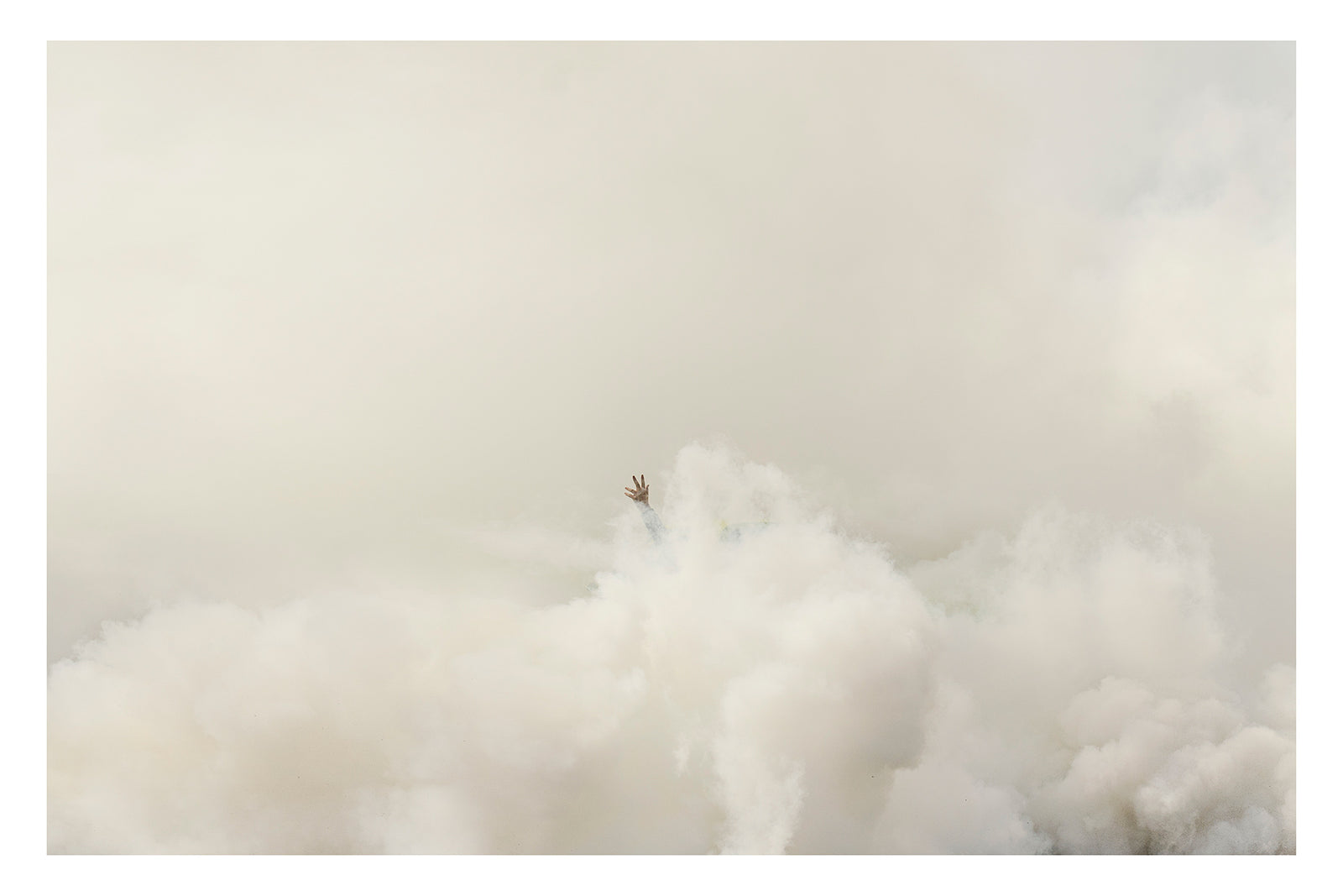 A single human hand reaches up from a vast rolling sea of white tyre smoke, fingers splayed open against a pale hazy sky, the rest of the body completely swallowed by the billowing cloud below. The minimalist composition and vast expanse of white smoke give the image an almost surreal, painterly quality. Fine art colour photographic print by Simon Davidson.
