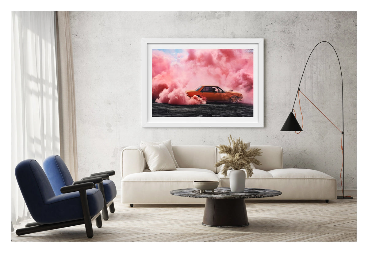 Framed in white, shown in a contemporary living space. We see an orange Ford XE Falcon sedan performs a burnout on heavily rubber marked black bitumen, surrounded by a cathedral of vivid pink burnout smoke that fills the sky above and wraps around the car on all sides. The driver is visible through the side window as pink smoke pours from the rear wheel arches, the warm orange body glowing against the soft pink haze with a glimpse of blue sky above. Fine art colour photographic print by Simon Davidson.