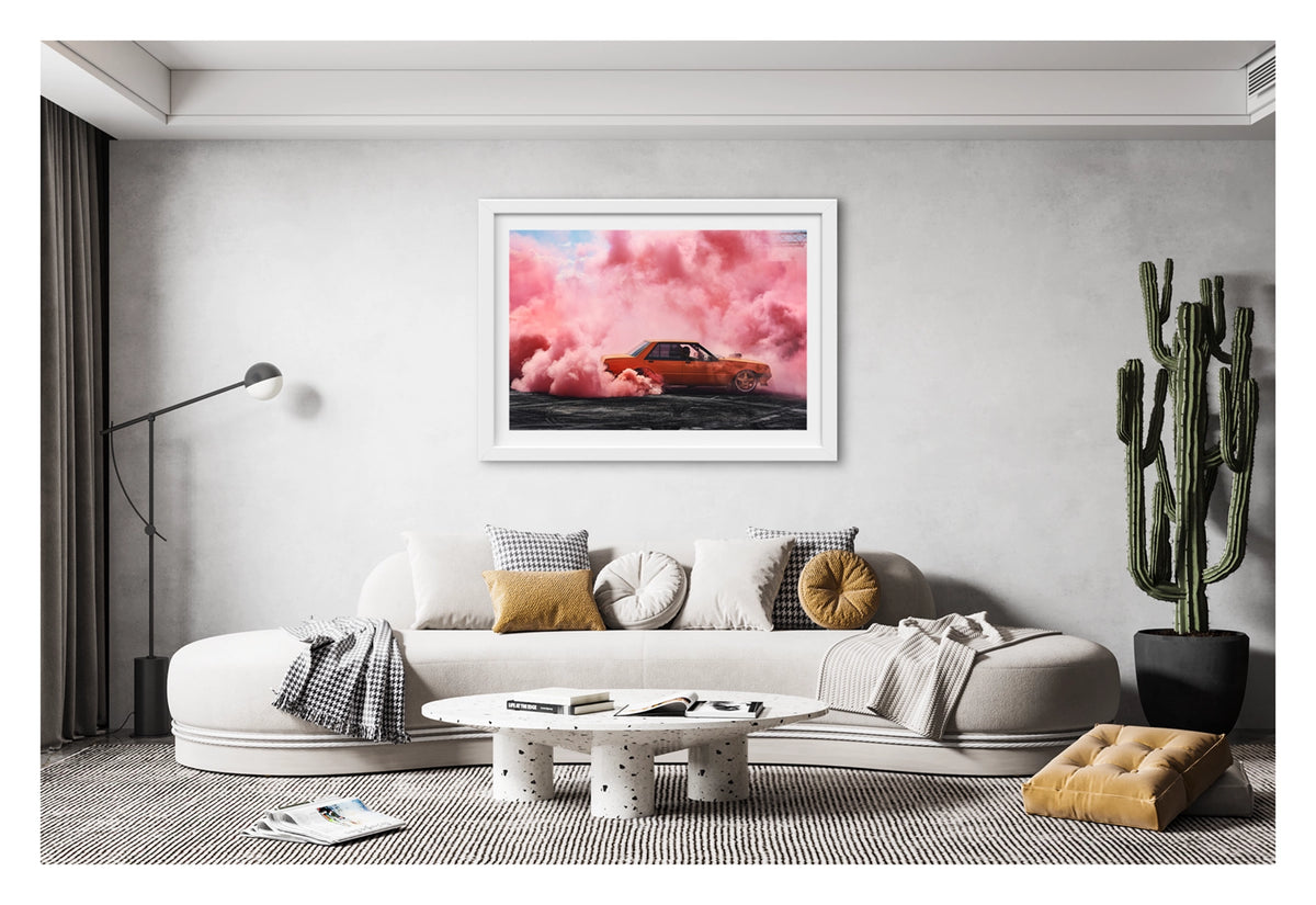 Framed in white, shown in a contemporary living space. We see an orange Ford XE Falcon sedan performs a burnout on heavily rubber marked black bitumen, surrounded by a cathedral of vivid pink burnout smoke that fills the sky above and wraps around the car on all sides. The driver is visible through the side window as pink smoke pours from the rear wheel arches, the warm orange body glowing against the soft pink haze with a glimpse of blue sky above. Fine art colour photographic print by Simon Davidson.