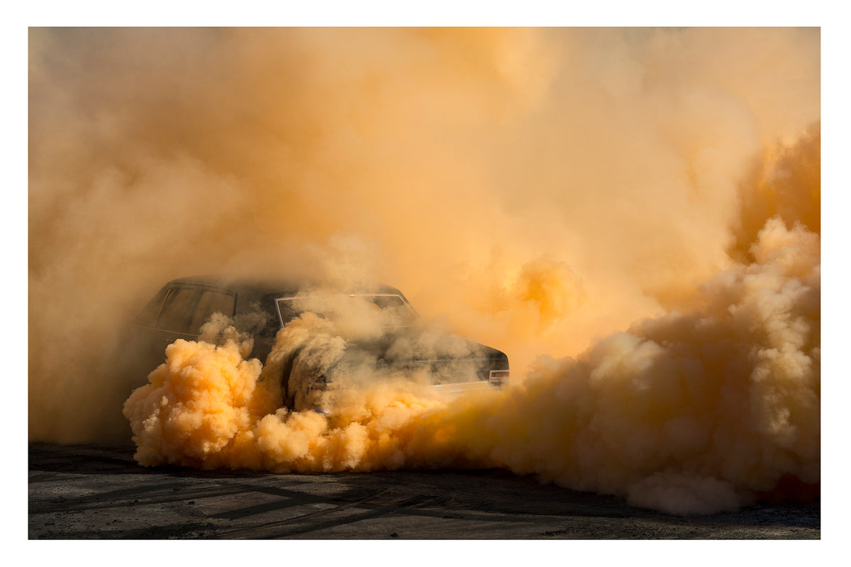 A black Holden Holden sedan is almost near consumed by a towering cloud of deep amber and orange tyre smoke that fills the entire frame, the roofline and windscreen barely visible through the churning golden haze. The warm orange smoke billows dramatically in every direction across the marked black bitumen, giving the scene an almost volcanic intensity. Fine art colour photographic print by Simon Davidson.