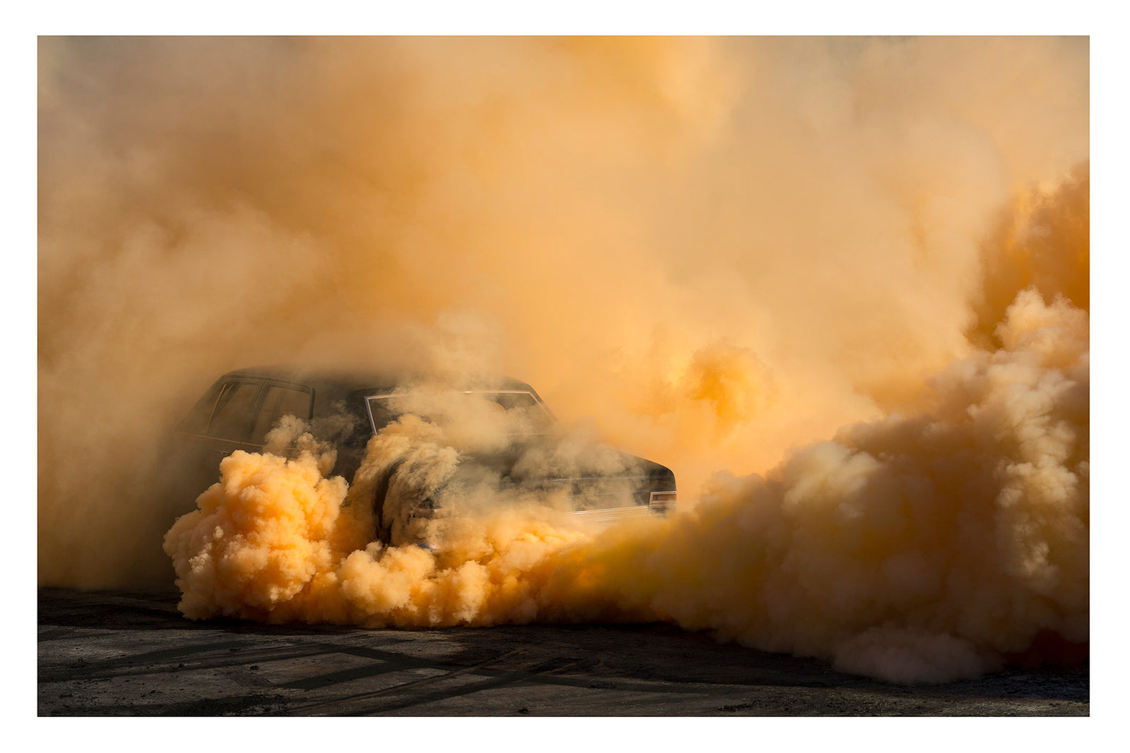 A black Holden Holden sedan is almost near consumed by a towering cloud of deep amber and orange tyre smoke that fills the entire frame, the roofline and windscreen barely visible through the churning golden haze. The warm orange smoke billows dramatically in every direction across the marked black bitumen, giving the scene an almost volcanic intensity. Fine art colour photographic print by Simon Davidson.