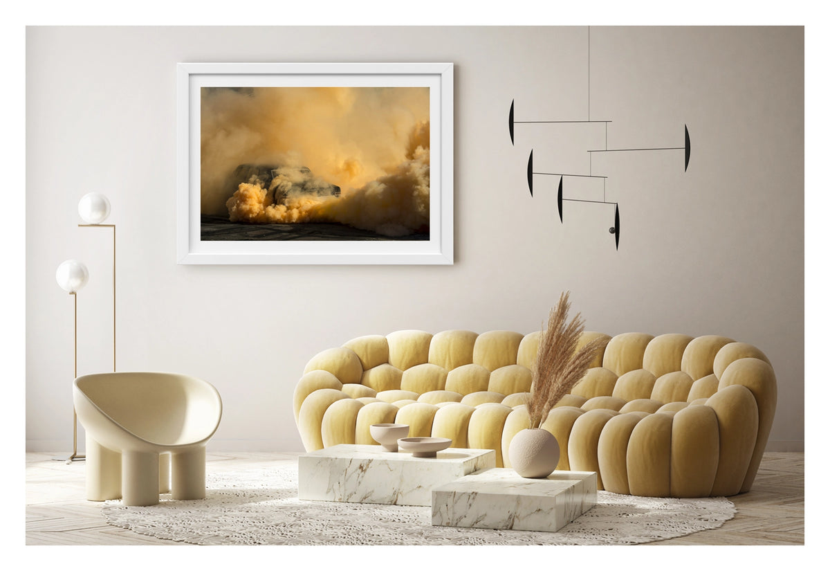 Framed in white, shown in a contemporary living space. A black Holden Holden sedan is almost near consumed by a towering cloud of deep amber and orange tyre smoke that fills the entire frame, the roofline and windscreen barely visible through the churning golden haze. The warm orange smoke billows dramatically in every direction across the marked black bitumen, giving the scene an almost volcanic intensity. Fine art colour photographic print by Simon Davidson.
