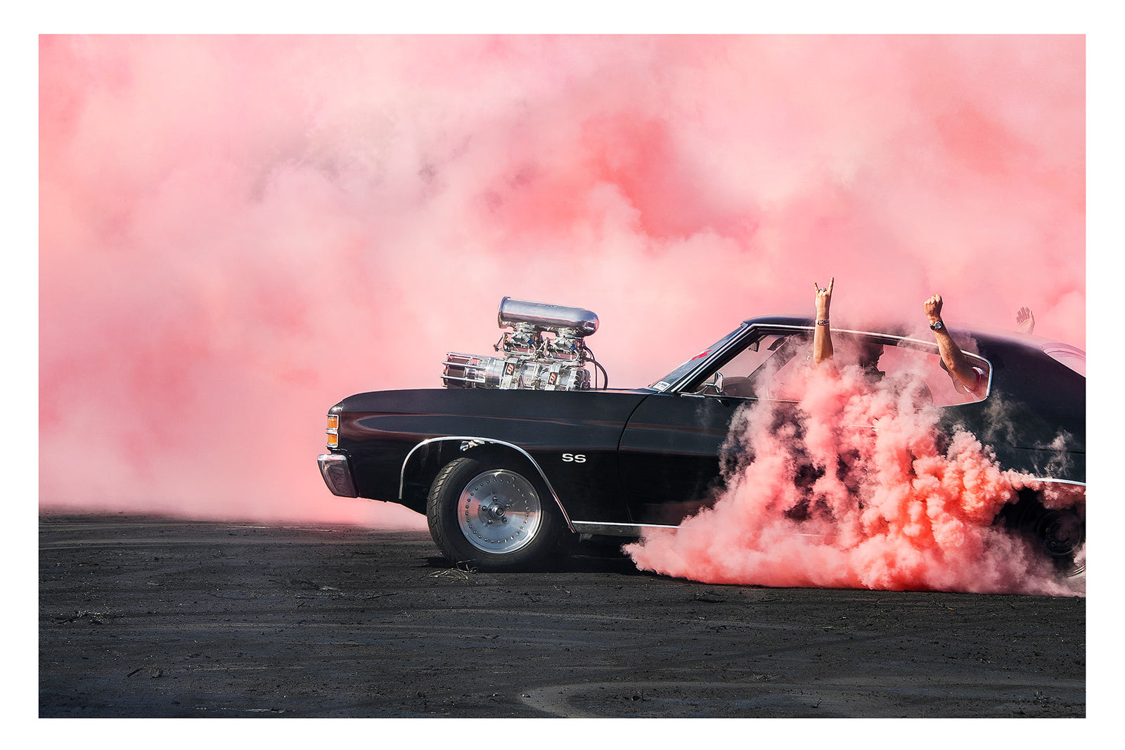 A black Chevrolet Chevelle SS with a massive polished supercharger protruding through the bonnet performs a burnout, engulfed in vivid pink tyre smoke that fills the entire background. The chrome blower and intake manifold gleam against the dark body as thick pink smoke pours from the under the car onto the tyre marked scorched black bitumen. Passengers'  arms out the windows in celebration. Fine art colour photographic print by Simon Davidson.