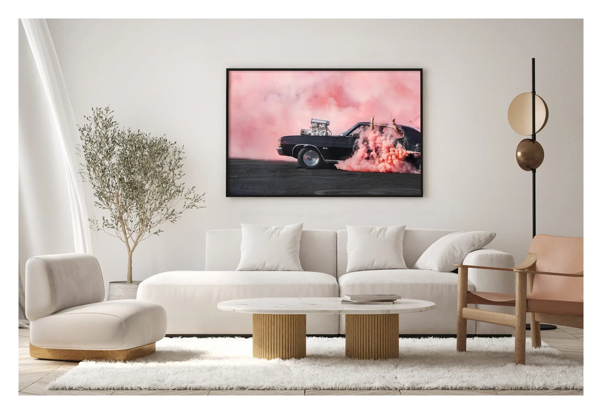 Framed in black. A black Chevrolet Chevelle SS with a massive polished supercharger protruding through the bonnet performs a burnout, engulfed in vivid pink tyre smoke that fills the entire background. The chrome blower and intake manifold gleam against the dark body as thick pink smoke pours from the under the car onto the tyre marked scorched black bitumen. Passengers'  arms out the windows in celebration. The print hangs in a contemporary lounge room. Fine art colour photographic print by Simon Davidson.