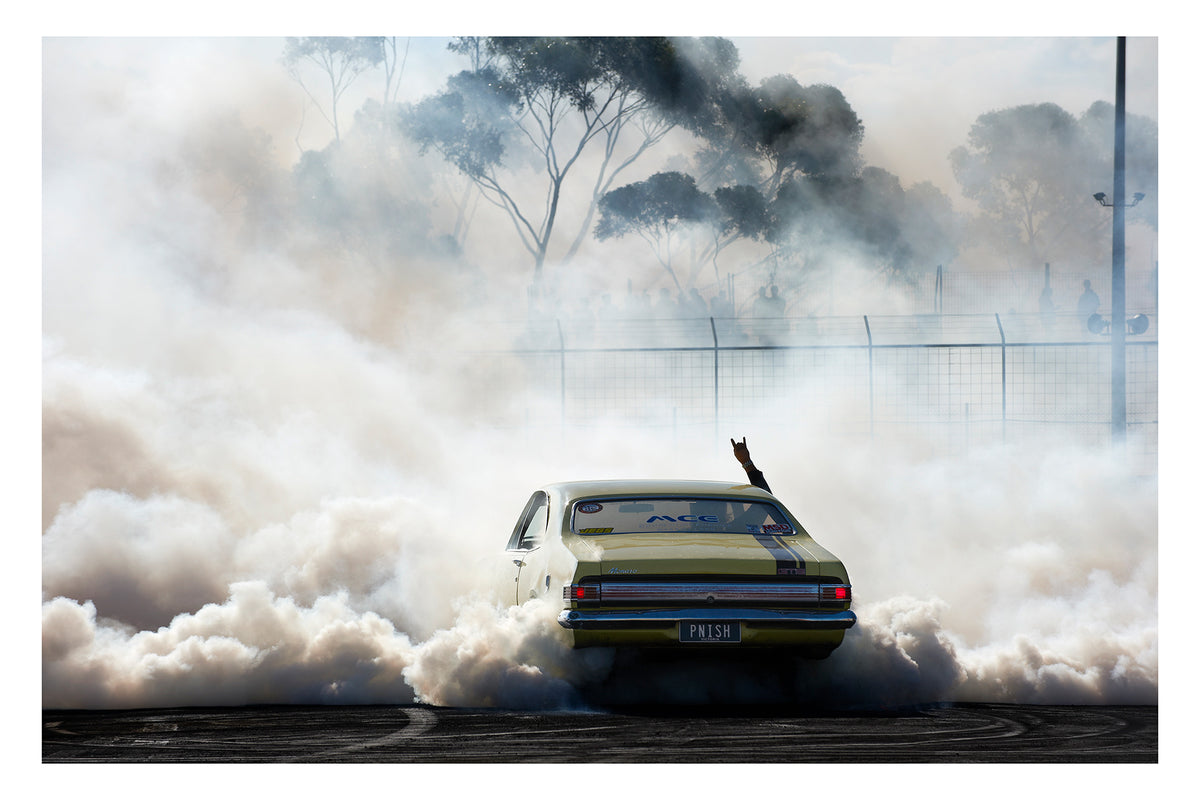 A yellow Holden HK GTS Monaro bearing the number plate PNISH performs a burnout at a competition, engulfed in a massive billowing cloud of white tyre smoke as a driver's arm reaches through the window throwing devil horns above the roofline. Australian eucalyptus trees ghost through the haze above a wire security fence in the background as the smoke rolls across the rubber marked bitumen surface in every direction. Fine art colour photographic print by Simon Davidson.