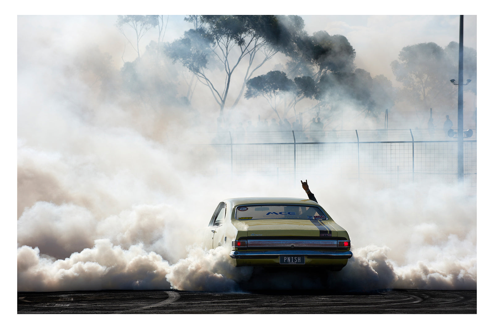 A yellow Holden HK GTS Monaro bearing the number plate PNISH performs a burnout at a competition, engulfed in a massive billowing cloud of white tyre smoke as a driver's arm reaches through the window throwing devil horns above the roofline. Australian eucalyptus trees ghost through the haze above a wire security fence in the background as the smoke rolls across the rubber marked bitumen surface in every direction. Fine art colour photographic print by Simon Davidson.