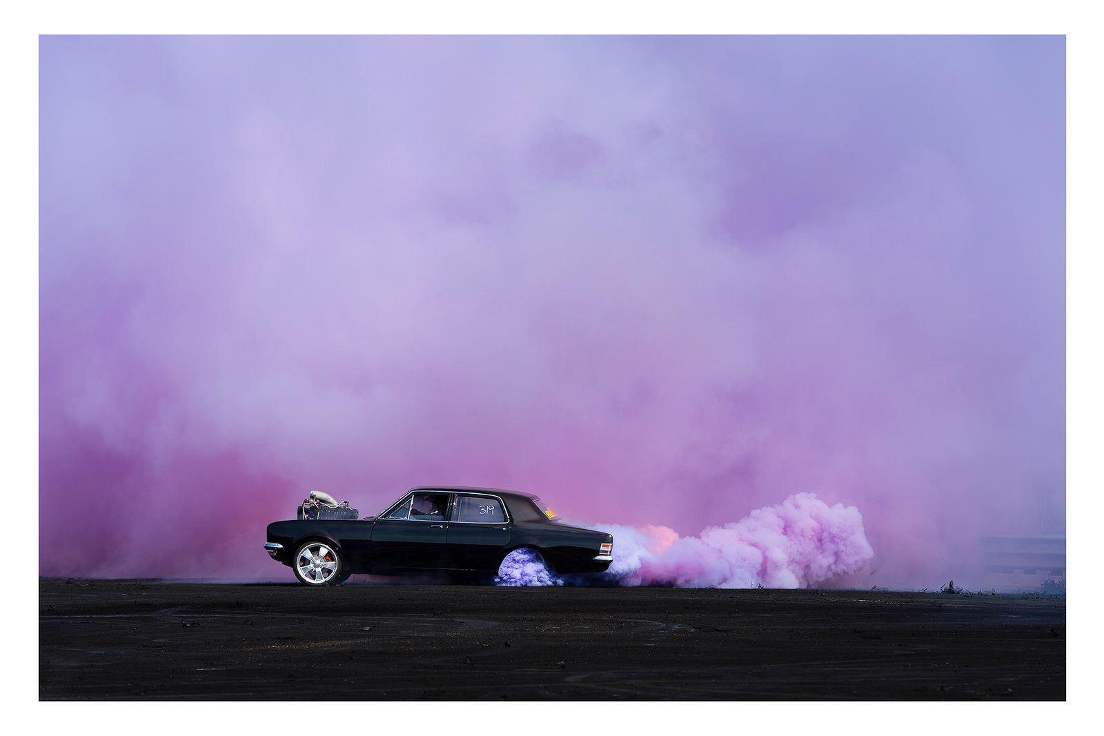 A black Holden chrome bumper sedan with an exposed supercharger out the bonnet performs a burnout on a vast black bitumen surface, trailing a long rolling cloud of billowing purple tyre smoke that sweeps across the right side of the frame and fills the entire sky behind. The car sits small and sharp against the expansive purple haze, the scene feeling open and cinematic in its scale. Fine art colour photographic print by Simon Davidson.