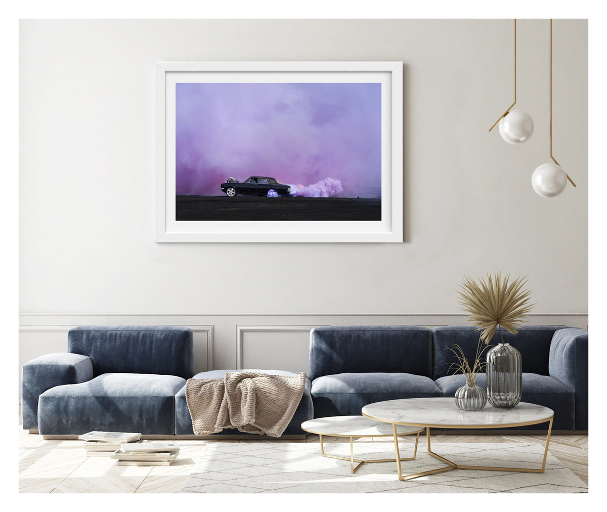 Framed in white, shown in a contemporary living space. A black Holden chrome bumper sedan with an exposed supercharger out the bonnet performs a burnout on a vast black bitumen surface, trailing a long rolling cloud of billowing purple tyre smoke that sweeps across the right side of the frame and fills the entire sky behind. The car sits small and sharp against the expansive purple haze, the scene feeling open and cinematic in its scale. Fine art colour photographic print by Simon Davidson.