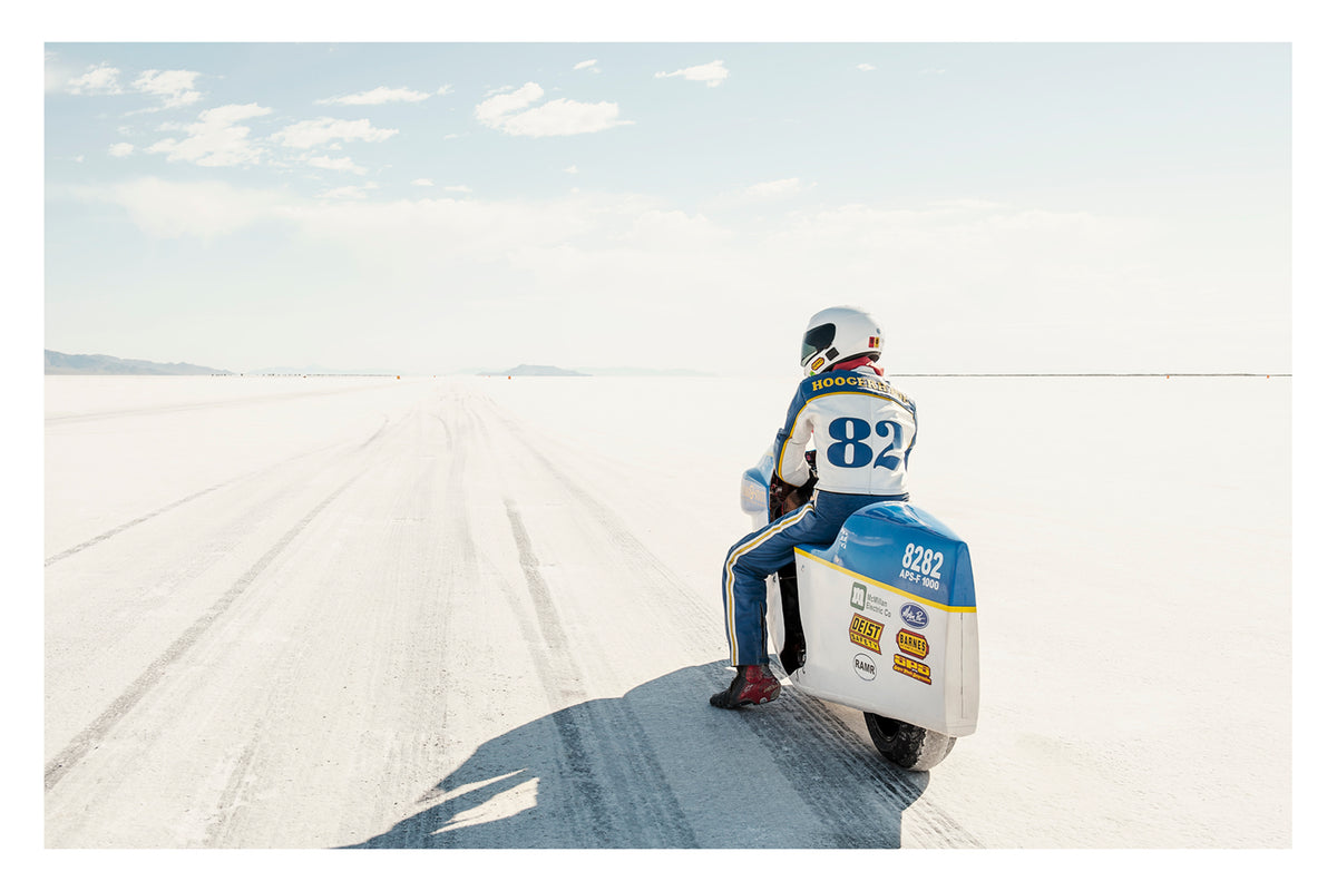 A land speed motorcycle racer wearing a blue and yellow race suit numbered 82 sits astride a white streamlined motorcycle numbered 8282 on the Bonneville Salt Flats, facing away from the camera toward the long straight track that stretches to the distant mountains and pale horizon. Tyre tracks score the white salt surface at the start line as a soft blue sky with scattered clouds completes the vast open scene. Fine art colour photographic print by Simon Davidson.