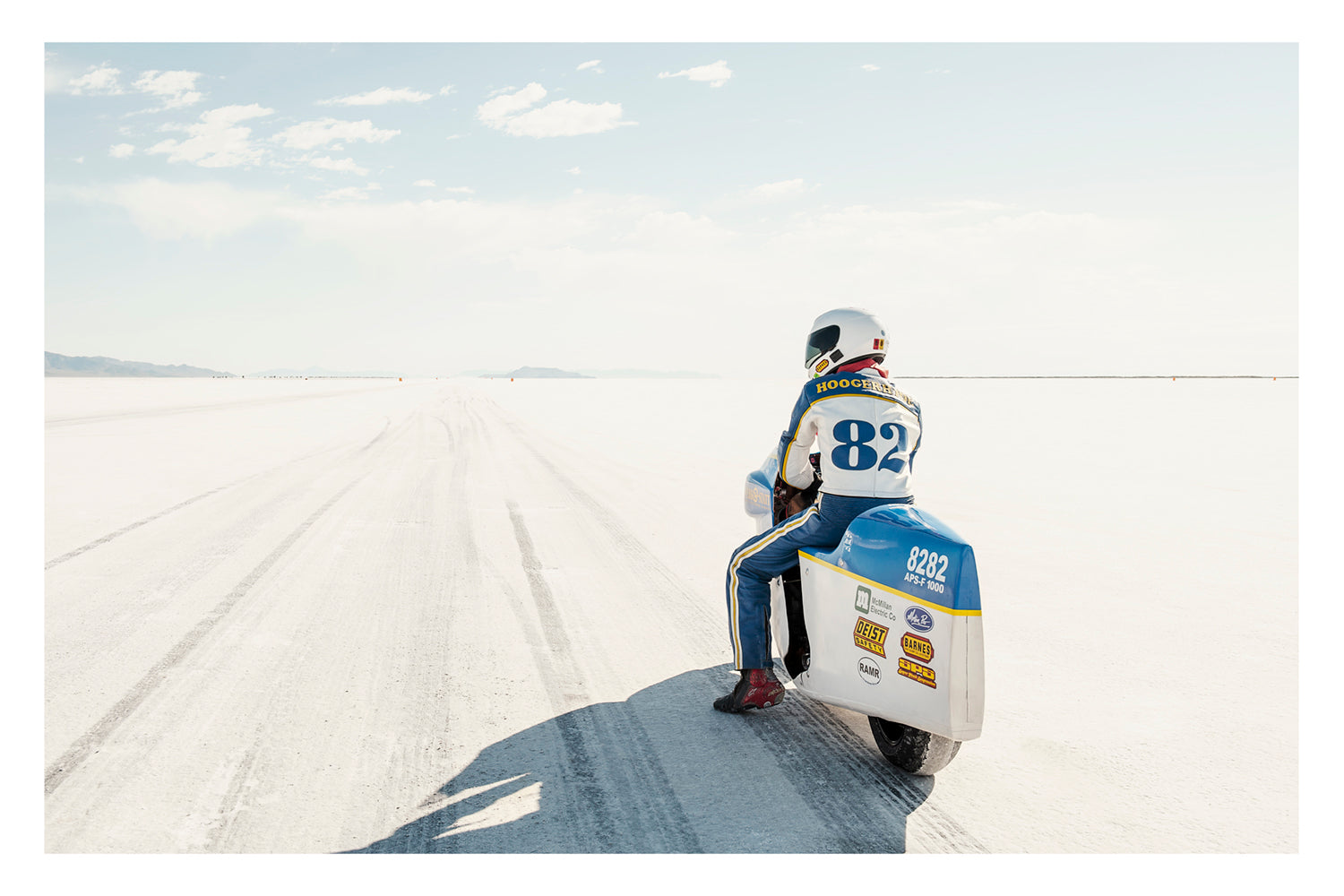 A land speed motorcycle racer wearing a blue and yellow race suit numbered 82 sits astride a white streamlined motorcycle numbered 8282 on the Bonneville Salt Flats, facing away from the camera toward the long straight track that stretches to the distant mountains and pale horizon. Tyre tracks score the white salt surface at the start line as a soft blue sky with scattered clouds completes the vast open scene. Fine art colour photographic print by Simon Davidson.