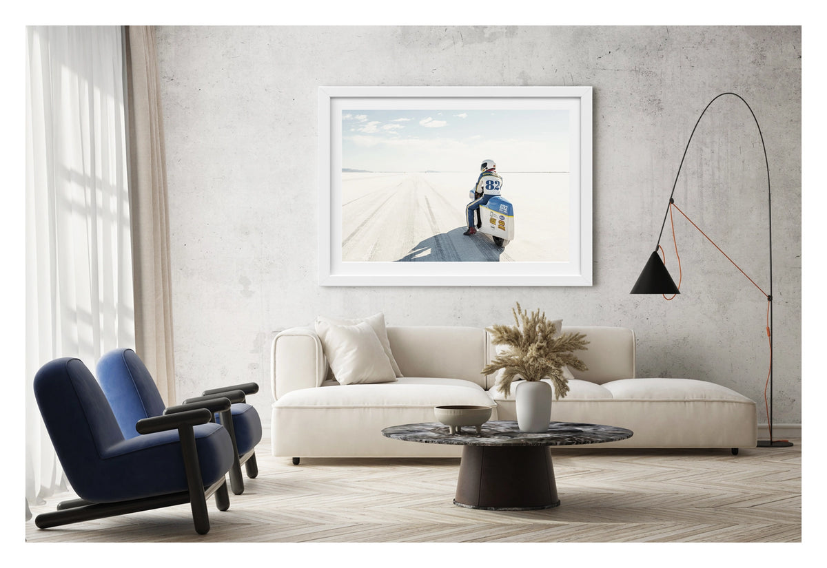Framed in white, shown in a modern space. A land speed motorcycle racer wearing a blue and yellow race suit numbered 82 sits astride a white streamlined motorcycle numbered 8282 on the Bonneville Salt Flats, facing away from the camera toward the long straight track that stretches to the distant mountains and pale horizon. Tyre tracks score the white salt surface at the start line as a soft blue sky with scattered clouds completes the vast open scene. Fine art colour photographic print by Simon Davidson.