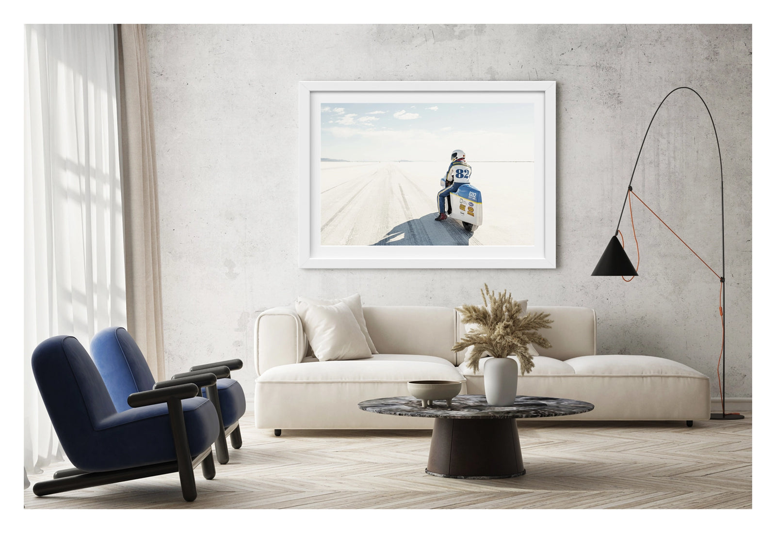 Framed in white, shown in a modern space. A land speed motorcycle racer wearing a blue and yellow race suit numbered 82 sits astride a white streamlined motorcycle numbered 8282 on the Bonneville Salt Flats, facing away from the camera toward the long straight track that stretches to the distant mountains and pale horizon. Tyre tracks score the white salt surface at the start line as a soft blue sky with scattered clouds completes the vast open scene. Fine art colour photographic print by Simon Davidson.