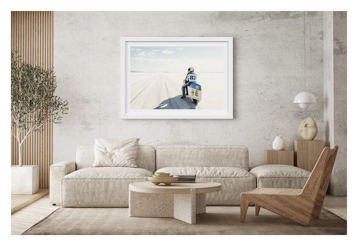 Framed in white, shown in a modern space. A land speed motorcycle racer wearing a blue and yellow race suit numbered 82 sits astride a white streamlined motorcycle numbered 8282 on the Bonneville Salt Flats, facing away from the camera toward the long straight track that stretches to the distant mountains and pale horizon. Tyre tracks score the white salt surface at the start line as a soft blue sky with scattered clouds completes the vast open scene. Fine art colour photographic print by Simon Davidson.