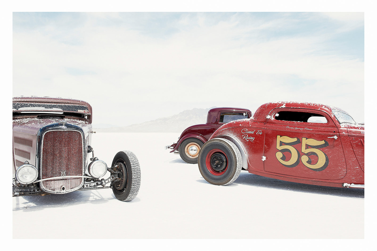 Two salt encrusted Rolling Bones hot rods sit on the blinding white surface of the Bonneville Salt Flats, their weathered bodies dusted with salt crystal residue. with a third maroon coupe visible between them in the middle distance. A pale mountain range dissolves into the white horizon behind them. Fine art colour photographic print by Simon Davidson.