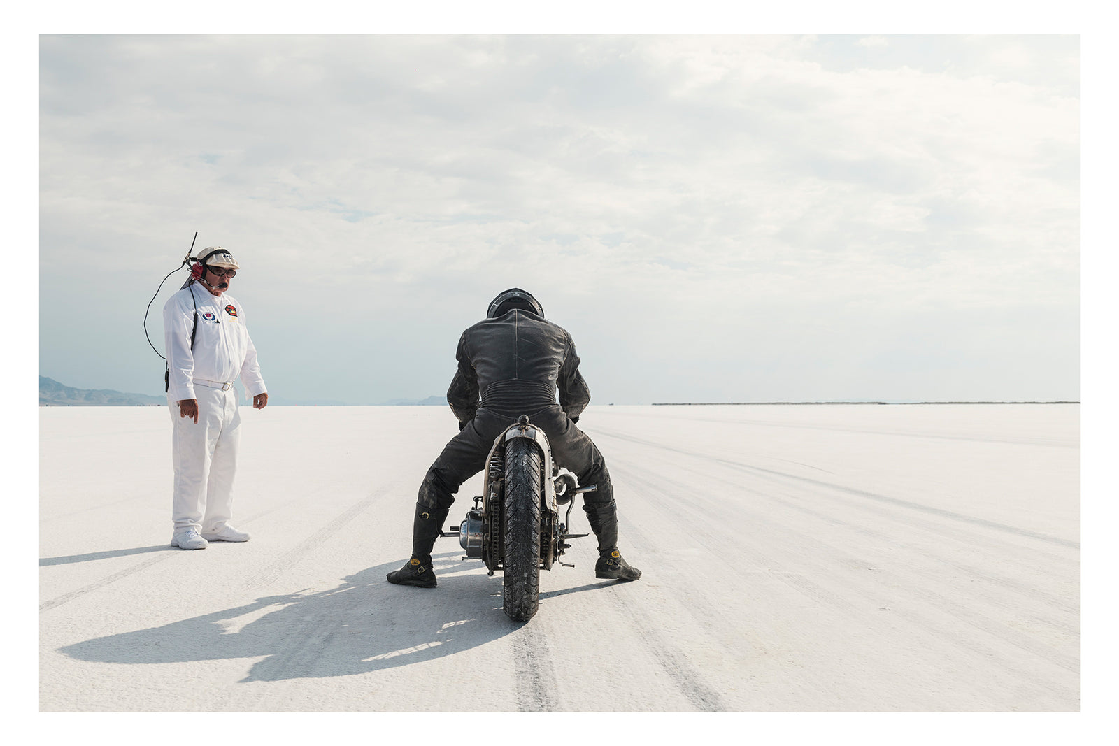 Motorcycle racer Shinya Kimura in black leathers crouches low over his vintage custom motorcycle on the Bonneville Salt Flats, poised at the start of a run as tyre tracks stretch toward the vanishing point ahead. A race official in a white jumpsuit and radio headset stands to the left watching on, the vast white salt and pale overcast sky dissolving into a seamless horizon in the distance. Fine art colour photographic print by Simon Davidson.