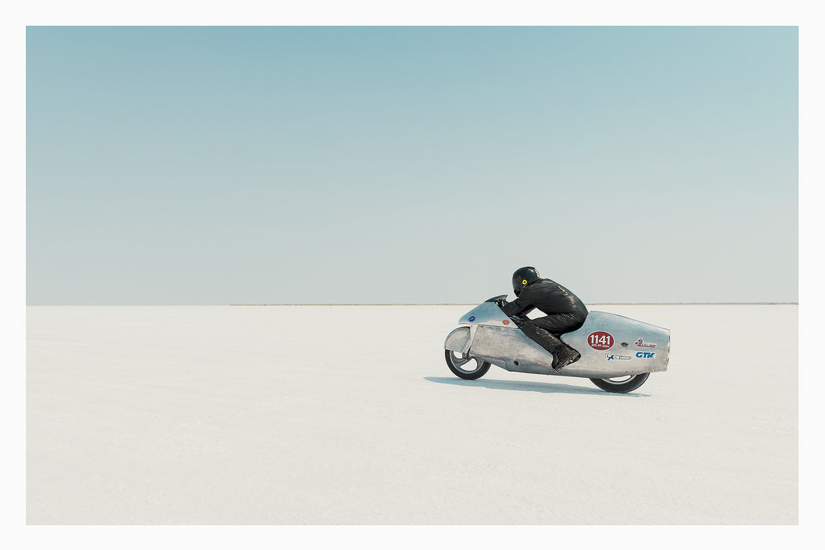 Clad in black leather, a rider crouches low over a bare metal aluminium streamliner motorcycle numbered 1141 as it races across the vast white expanse of the bonneville  salt flats The rider and machine are small but in striking form against the pale horizon and soft blue sky above. The hand beaten metal bike and minimal silhouette give the scene a timeless, sculptural quality as it pushes across the featureless white surface. Fine art colour photographic print by Simon Davidson.