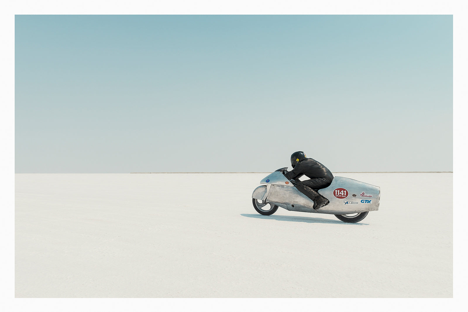 Clad in black leather, a rider crouches low over a bare metal aluminium streamliner motorcycle numbered 1141 as it races across the vast white expanse of the bonneville  salt flats The rider and machine are small but in striking form against the pale horizon and soft blue sky above. The hand beaten metal bike and minimal silhouette give the scene a timeless, sculptural quality as it pushes across the featureless white surface. Fine art colour photographic print by Simon Davidson.