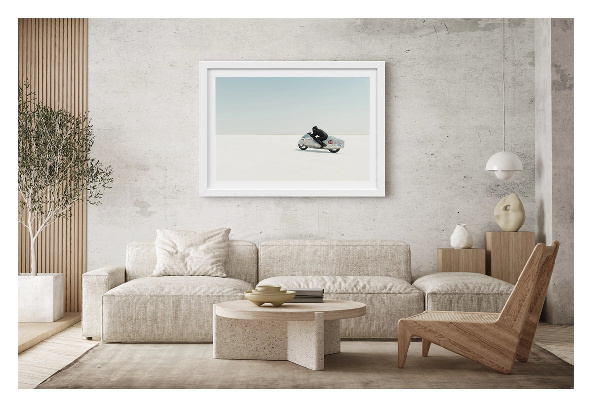 Framed in white, shown in a contemporary living space is a black leather clad rider crouching low over a bare metal aluminium streamliner motorcycle numbered 1141 as it races across the vast white expanse of the bonneville  salt flats The rider and machine are small but in striking form against the pale horizon and soft blue sky above. The hand beaten metal bike and minimal silhouette give the scene a timeless, sculptural quality as it pushes across the featureless white surface. Fine art colour photograph.
