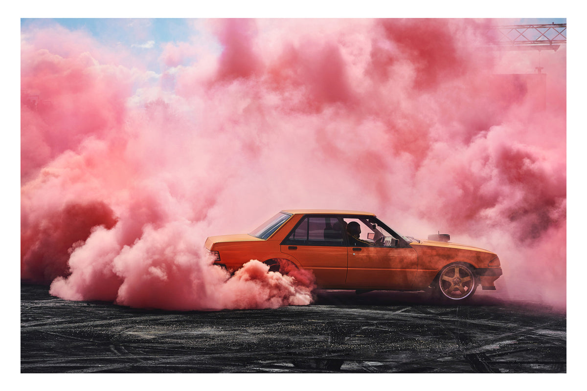 An orange Ford XE Falcon sedan performs a burnout on heavily rubber marked black bitumen, surrounded by a cathedral of vivid pink burnout smoke that fills the sky above and wraps around the car on all sides. The driver is visible through the side window as pink smoke pours from the rear wheel arches, the warm orange body glowing against the soft pink haze with a glimpse of blue sky above. Fine art colour photographic print by Simon Davidson.
