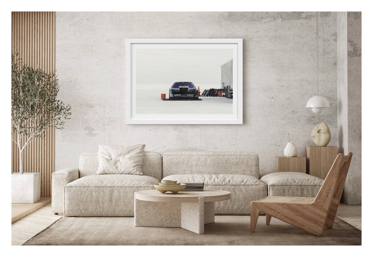 Framed in white and shown in a contemporary space. A black Chevrolet Corvette land speed racing car sits low on a ground sheet in the pits at Bonneville. Surrounded by the vast white expanse of the Salt Flats, faced directly toward the camera. Orange buckets, a row of spare tyres leaning against a trailer and an orange traffic cone are scattered around the pit area to the right, the whole scene dissolving into a featureless white horizon and sky. Fine art colour photographic print by Simon Davidson.