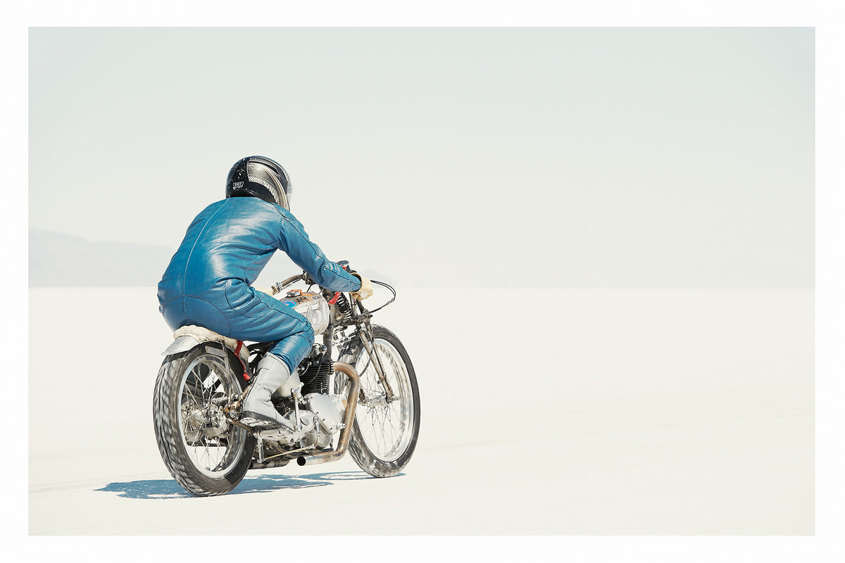 A lone motorcycle racer dressed in a blue leather suit and silver helmet rides a vintage Triumph, land speed racing motorcycle across the blinding white expanse of the Bonneville salt flats, the rider and bike reduced to a small solitary figure against an almost entirely white horizon and washed out sky. Fine art colour photographic print by Simon Davidson.