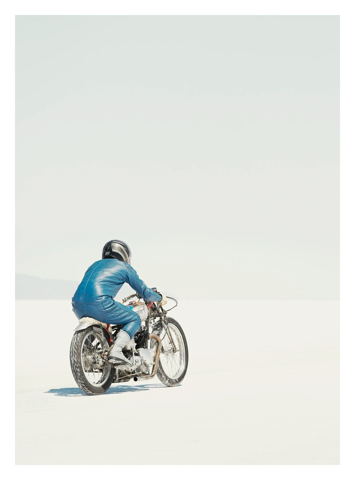 Captured in portrait orientation. A lone motorcycle racer dressed in a blue leather suit and silver helmet rides a vintage Triumph, land speed racing motorcycle across the blinding white expanse of the Bonneville salt flats, the rider and bike reduced to a small solitary figure against an almost entirely white horizon and pale sky. Fine art colour photographic print by Simon Davidson.