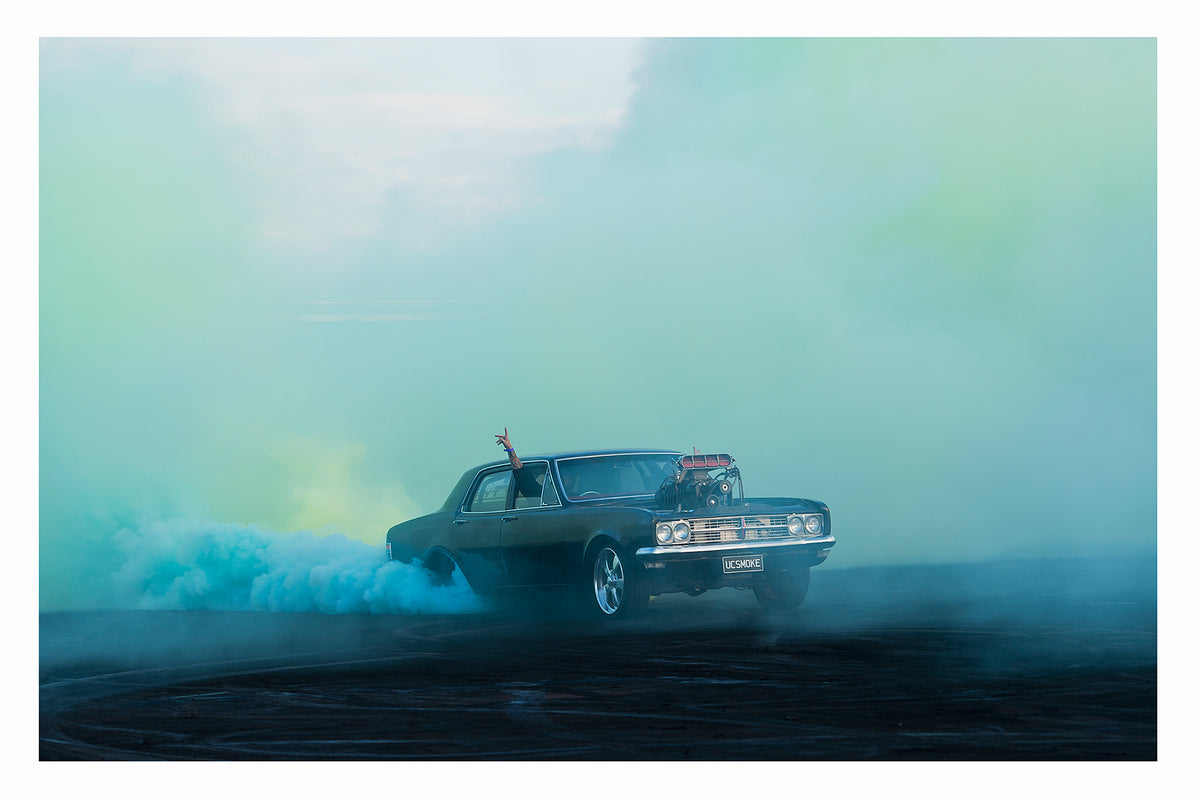 A black Holden HD sedan with a supercharger out the bonnet, bearing the number plate UCSMOKE, performs a burnout surrounded by vast aquamarine tyre smoke that bathes the entire scene in soft blue green light. A driver's arm reaches out the window above the roofline, as aqua smoke rolls low across the ground and the ethereal coloured haze fills the sky behind. Fine art colour photographic print by Simon Davidson.