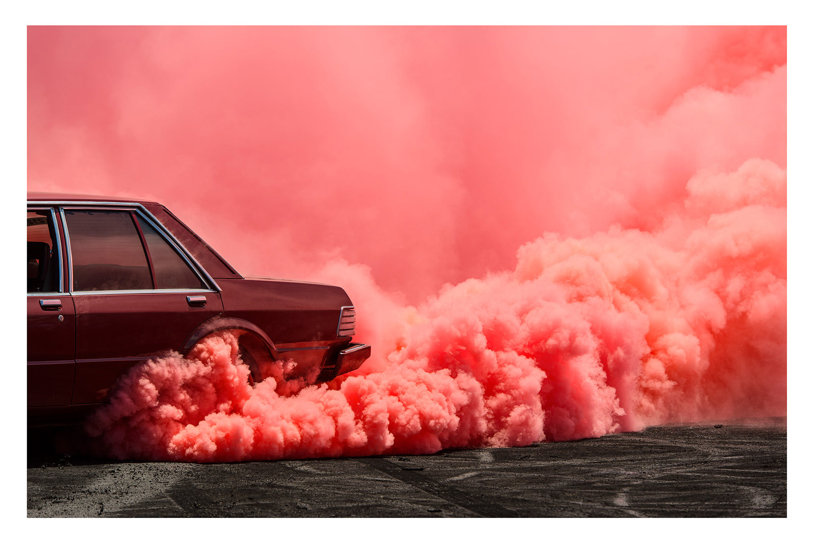 The visible rear lefthand quarter of a dark burgundy Ford Falcon sedan performs a burnout, a massive rolling cloud of vivid pink tyre smoke erupting from the rear wheel arch and sweeping across the full width of the frame. The deep red body of the car sits in rich tonal harmony with the warm pink smoke that floods the sky above and rolls heavily across the tyre marked burnout pad. Fine art colour photographic print by Simon Davidson.