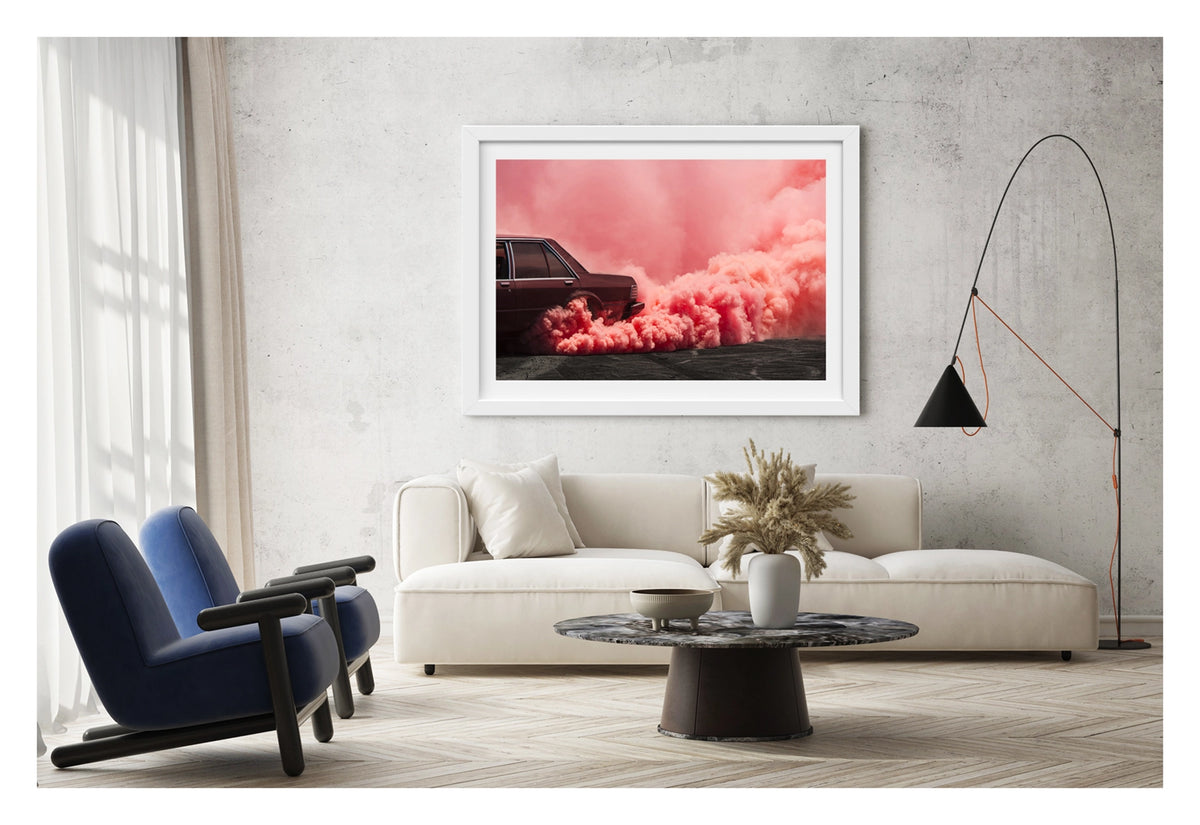 Framed in white, shown in a contemporary living space. The visible rear lefthand quarter of a dark burgundy Ford Falcon sedan is mid burnout, a massive rolling cloud of vivid pink tyre smoke erupting from the rear wheel arch and sweeping across the full width of the frame. The deep red body of the car sits in rich tonal harmony with the warm pink smoke that floods the sky above and rolls heavily across the tyre marked burnout pad. Fine art colour photographic print by Simon Davidson.